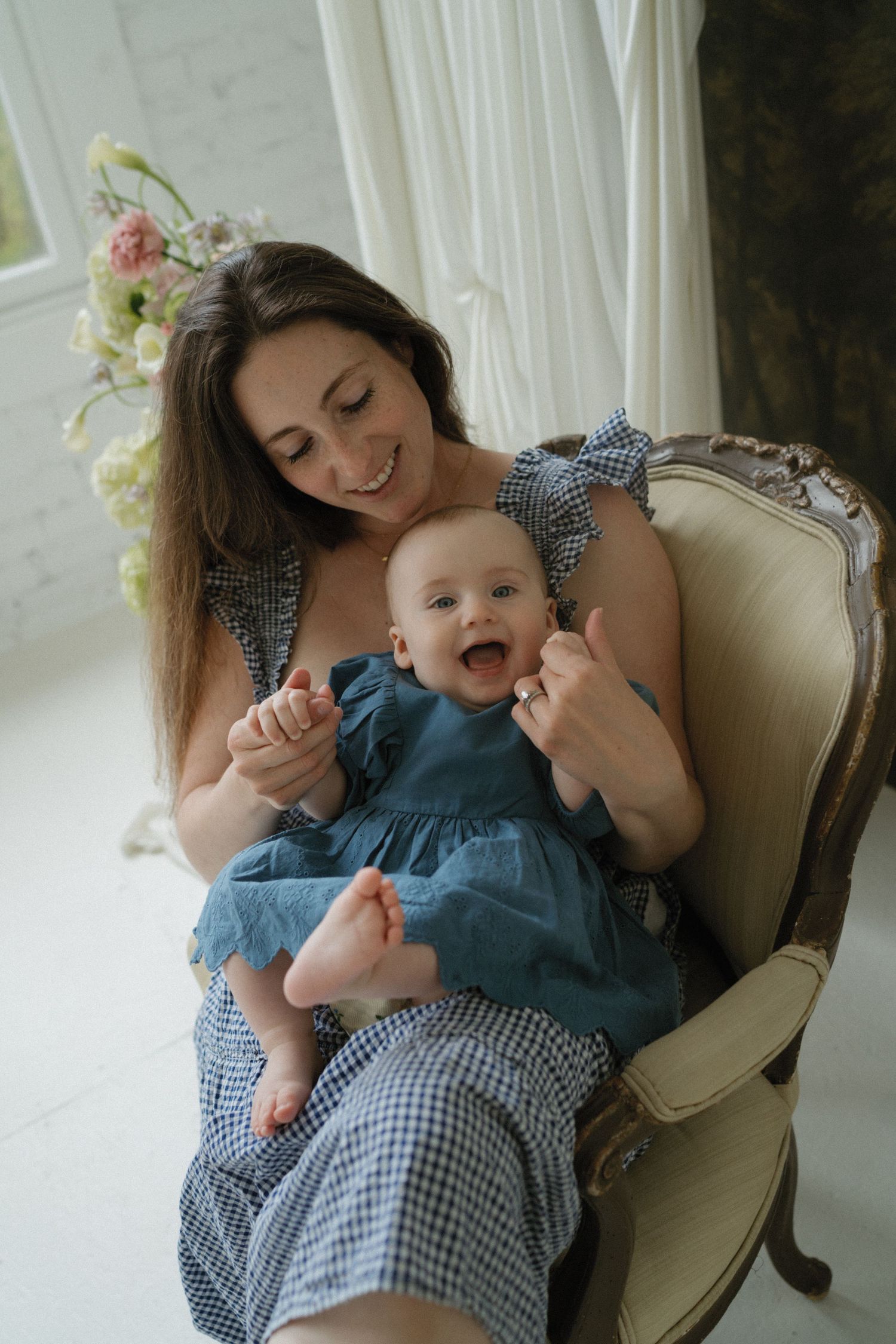 Smiling mother in blue dress holding happy baby girl in matching blue dress on vintage armchair.