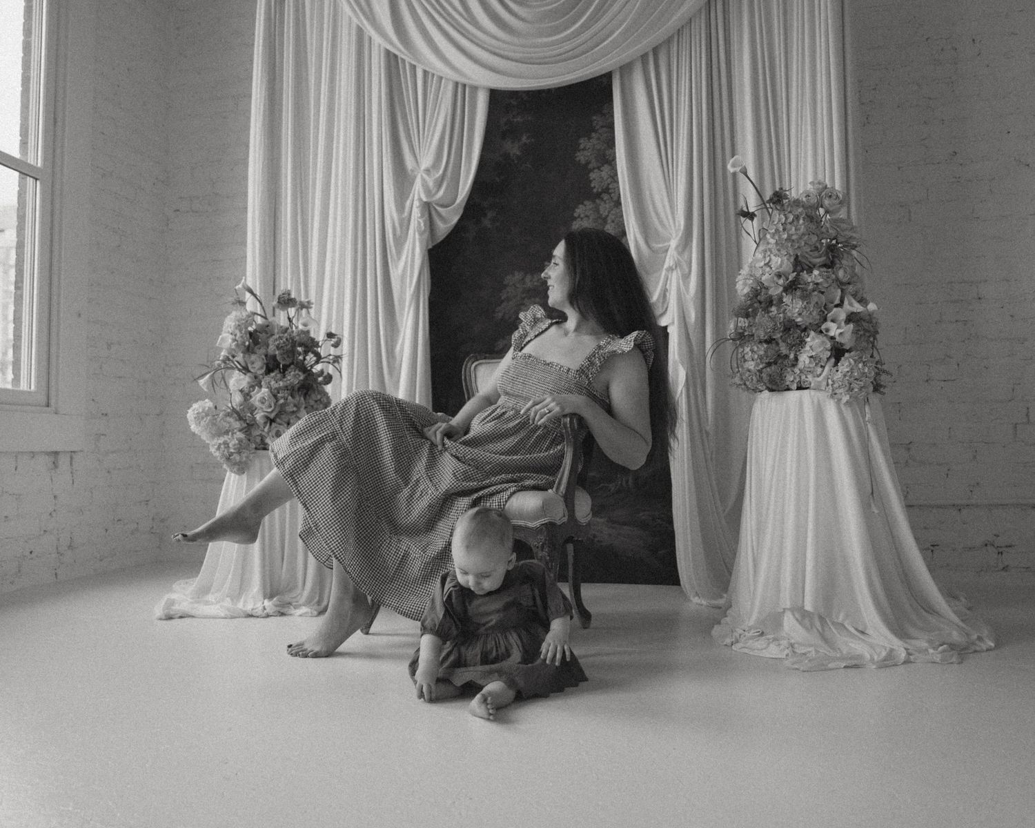 Black and white maternity photo with a pregnant woman in a flowy gown seated near draped curtains and floral arrangements.