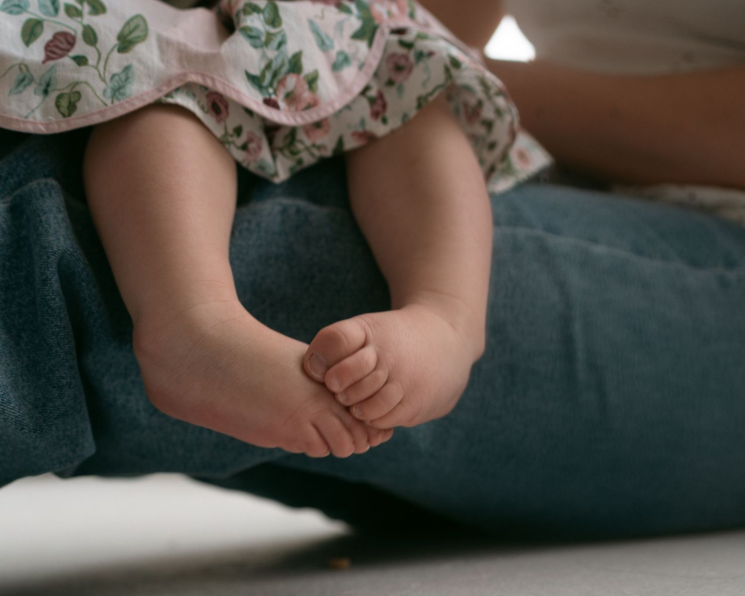 Close-up of a baby's tiny bare feet resting on an adult's denim-clad lap, wearing a floral dress.