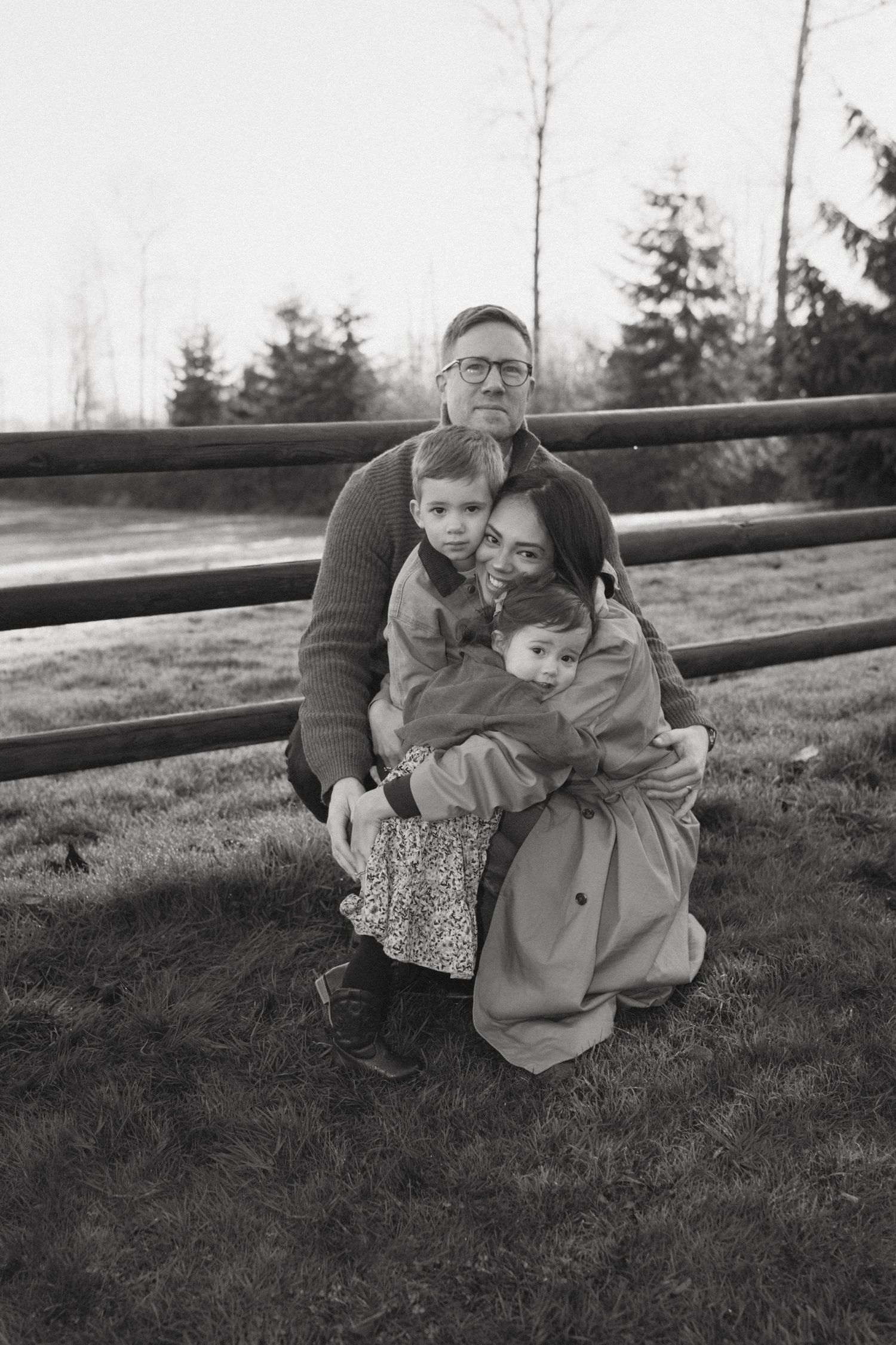 Black and white photo of family members sharing an affectionate embrace on grassy hillside near wooden fence.