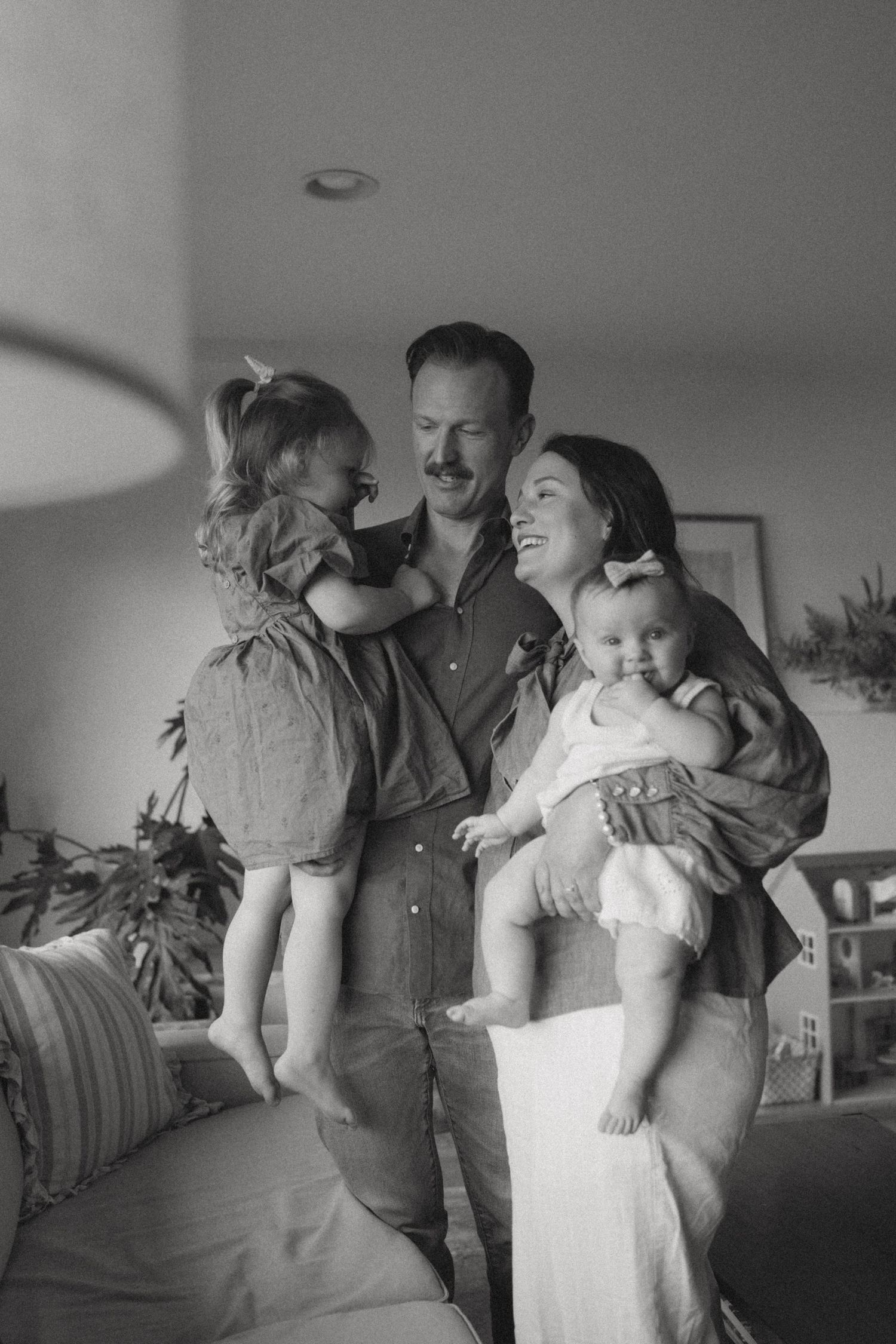 Black and white family portrait of smiling parents holding a toddler and baby in a cozy living room with a sofa and plants.