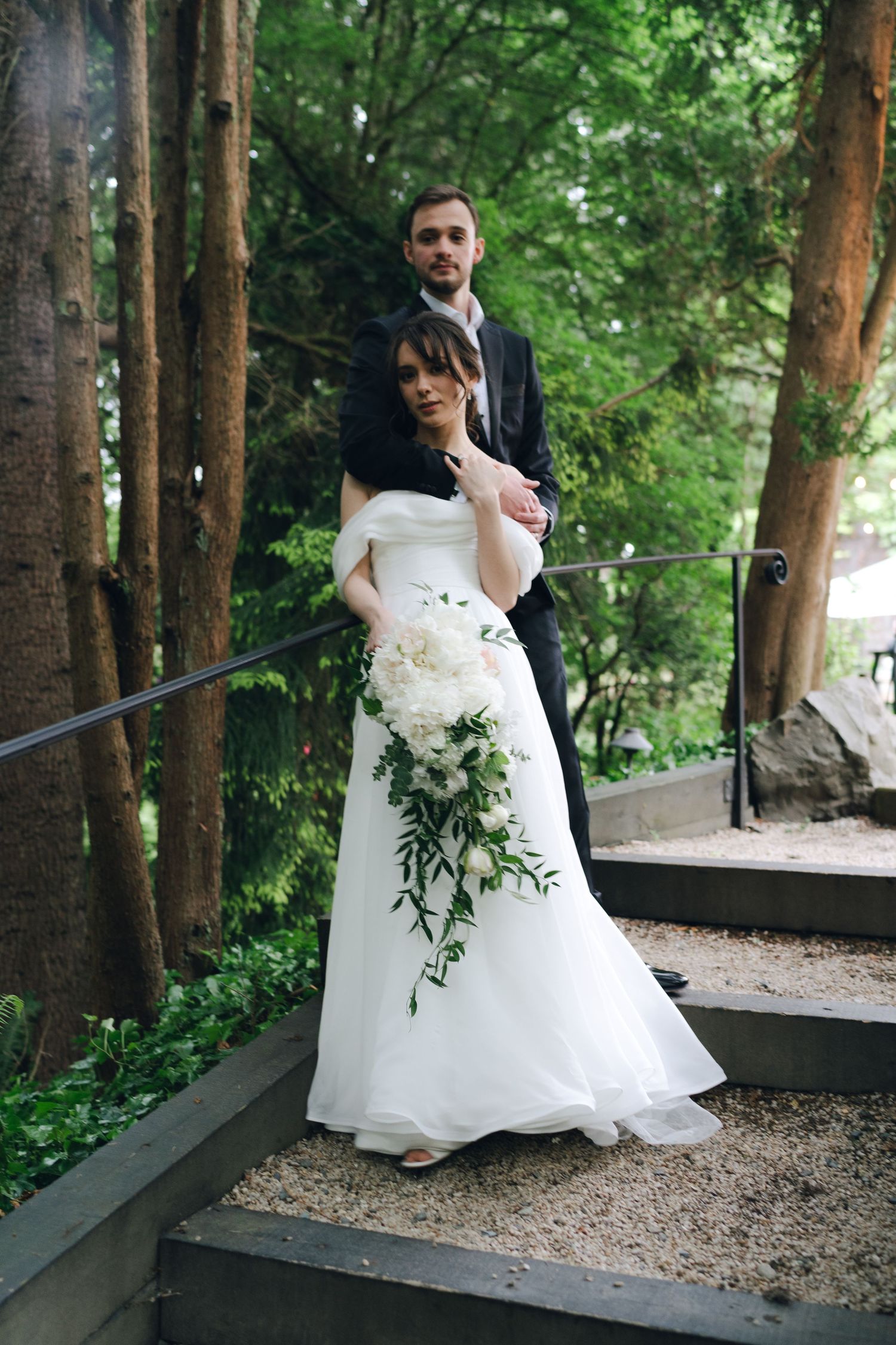 A couple in wedding attire poses along a rustic wooden railing in a wooded setting.