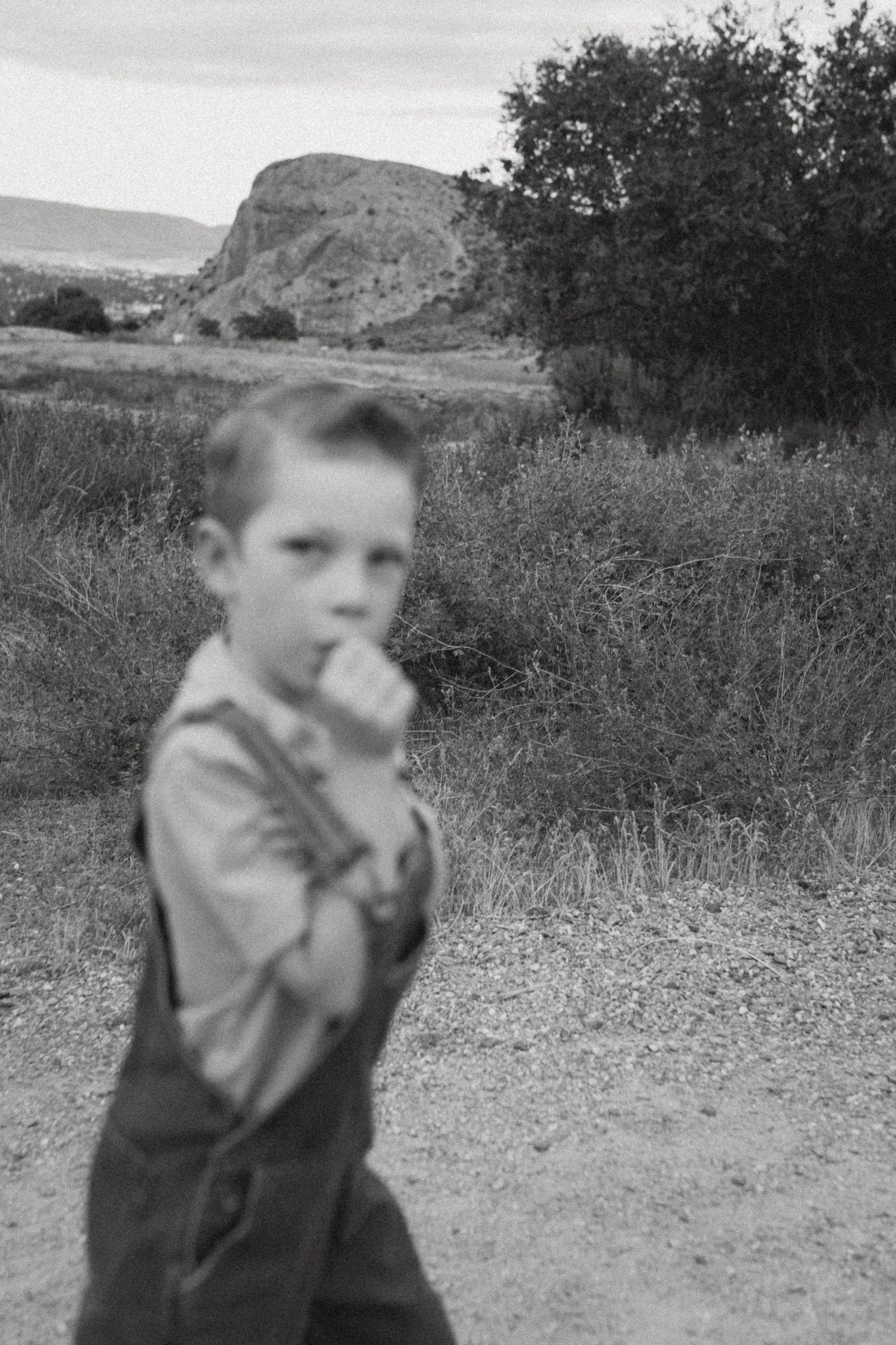 A black and white photo of a young boy in overalls with his fist near his mouth, standing on a dirt path before a rocky hill.