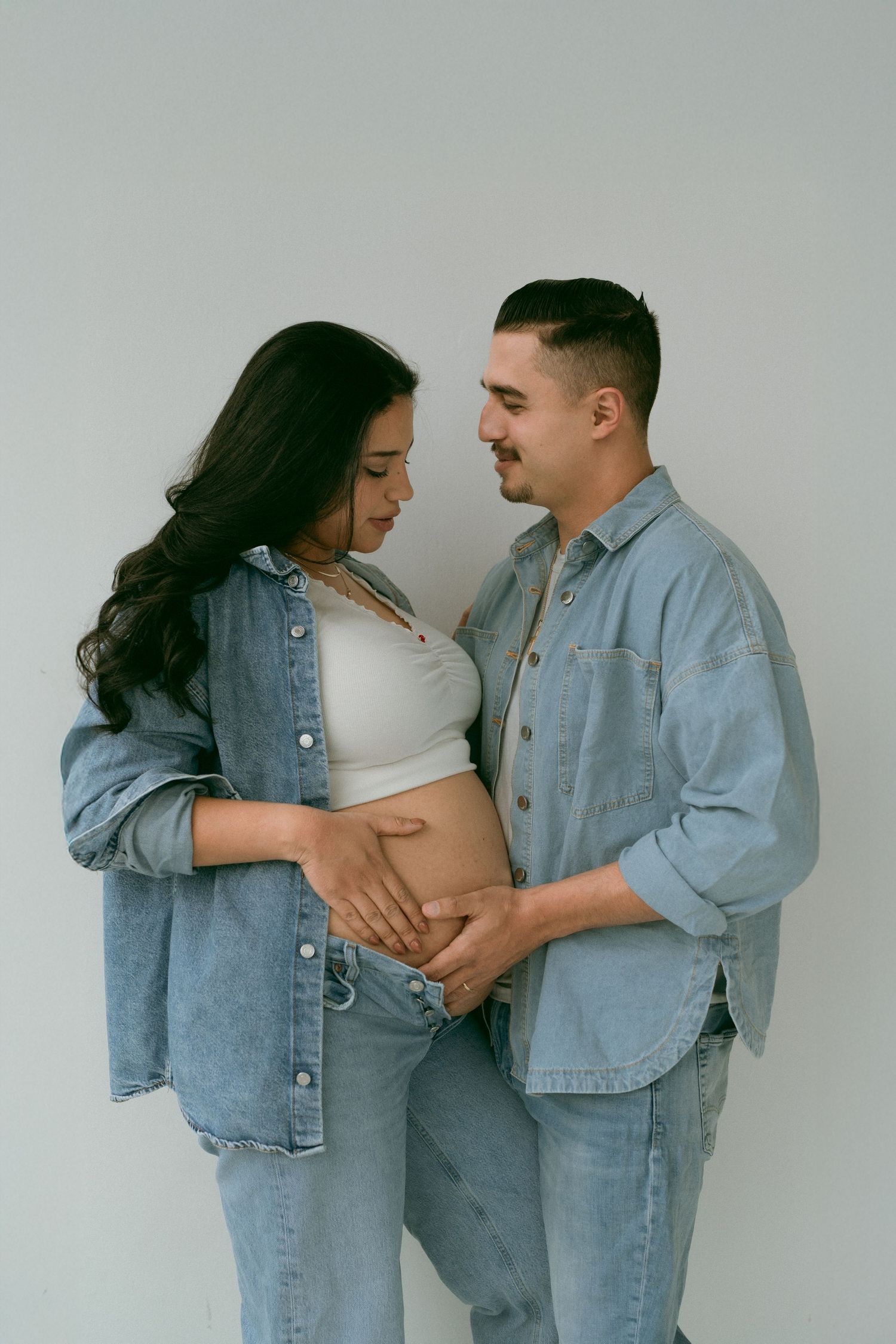 Couple wearing matching denim outfits embraces tenderly against white background in romantic photo series.