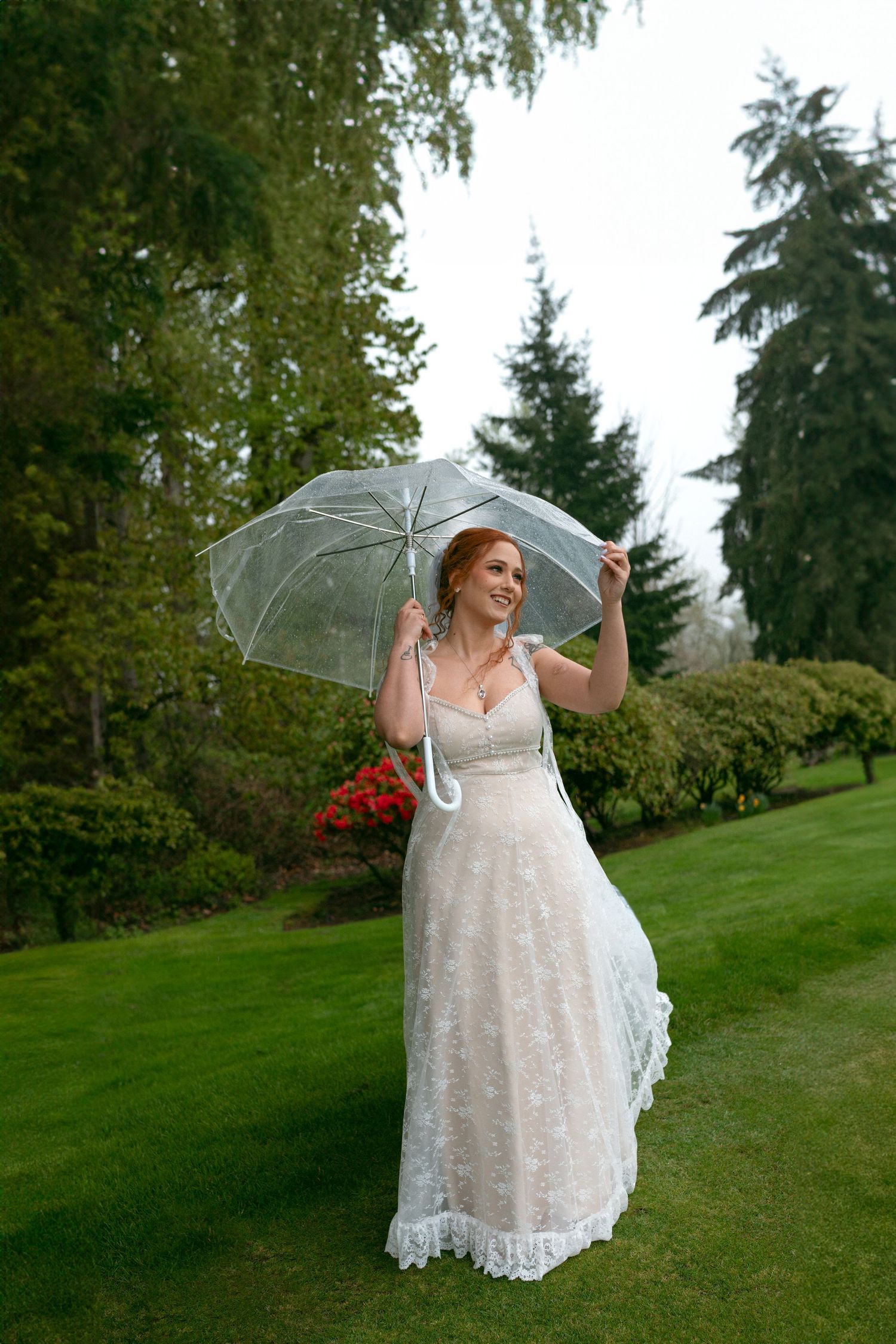 A person in a lace wedding dress poses joyfully with a clear umbrella on grassy hillside.