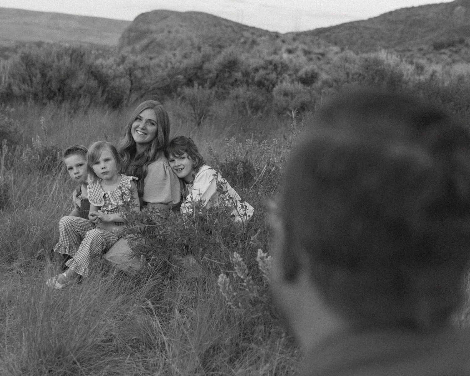 Black and white photo of a smiling mother and three young children sitting together in tall grass on a hillside as a photographer shoots.