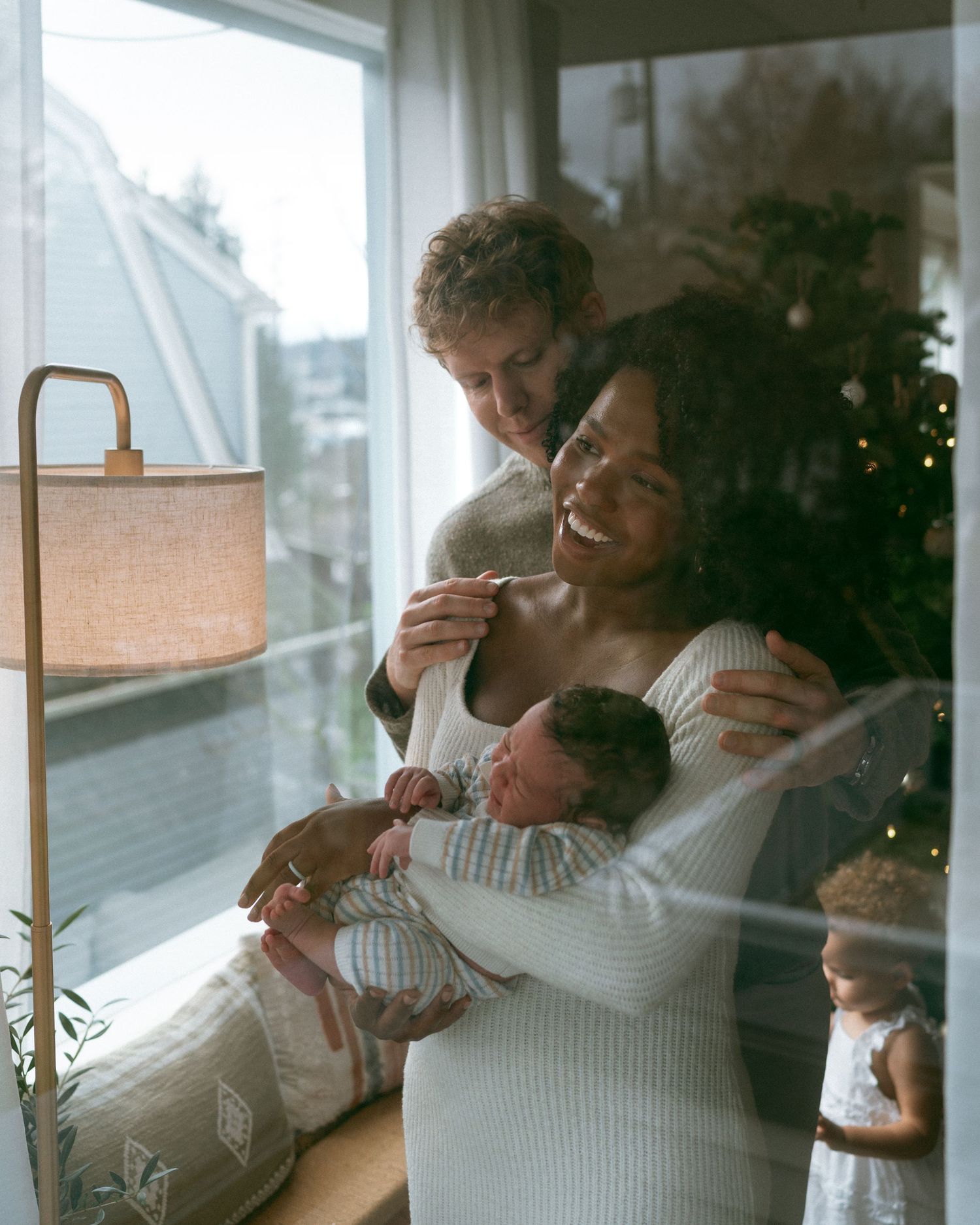Two people share an intimate embrace while sitting by a window in matching white sweaters.