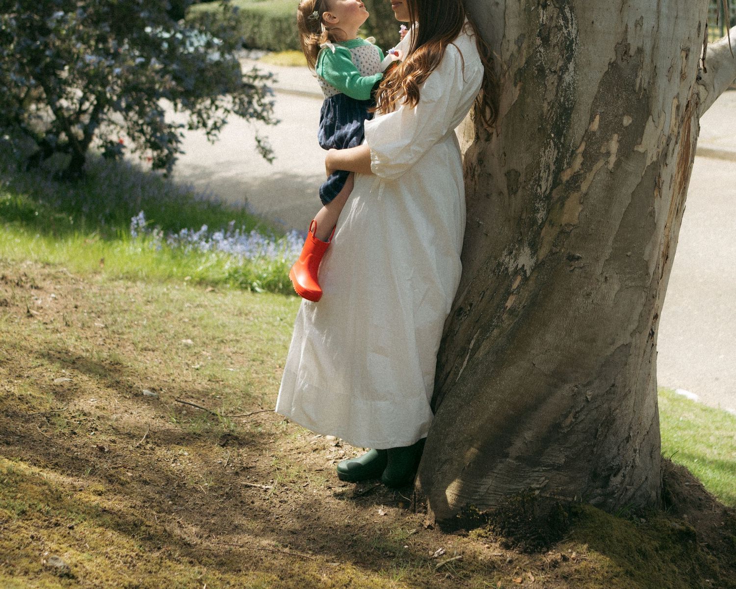 Woman in white dress holding a child beside a large tree trunk in a sunny park with blue flowers.