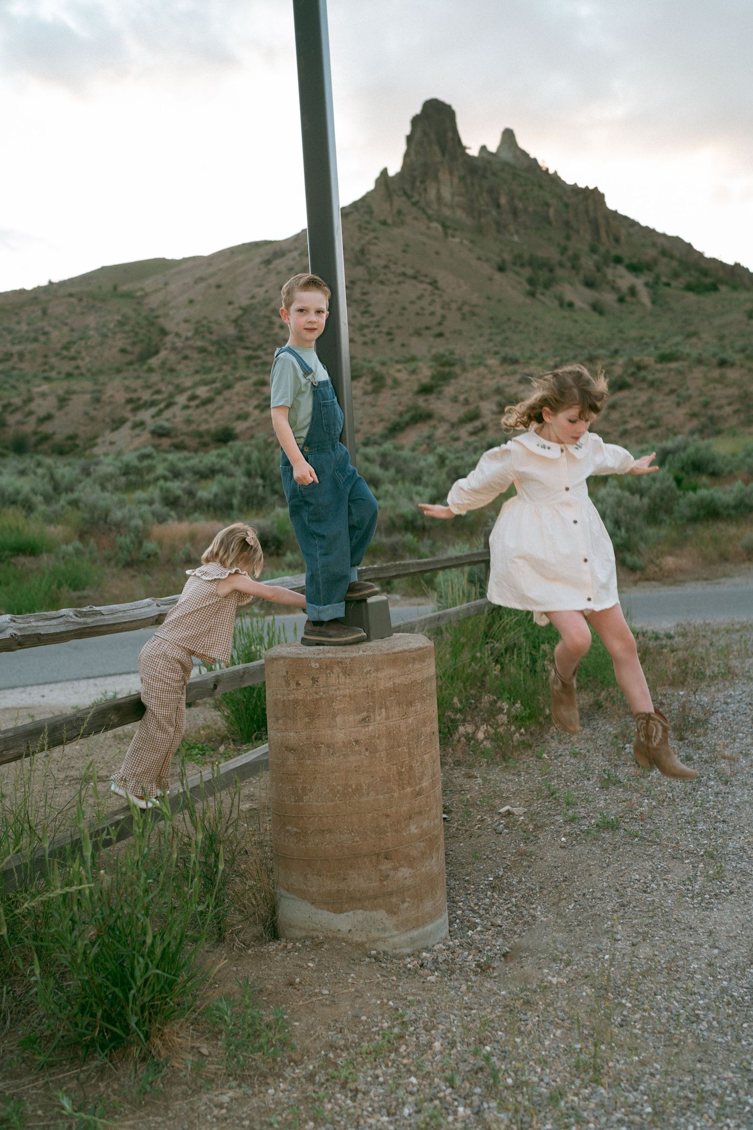 Three children in denim overalls and cowboy boots play on a wooden fence and concrete post near a rocky desert hillside at dusk.