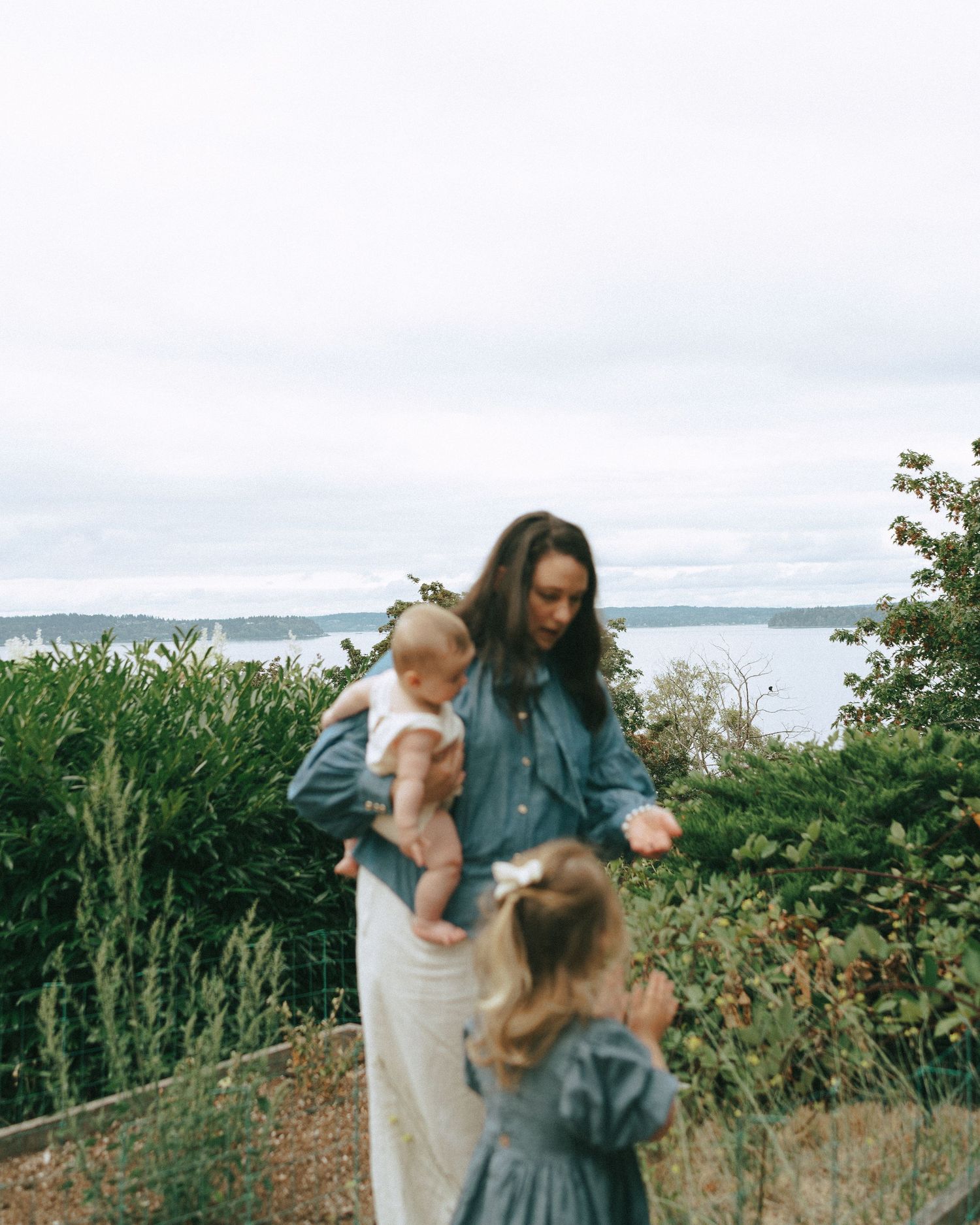 A woman in a denim shirt holds a baby while a toddler in a green dress stands nearby in a garden overlooking calm water on a cloudy day.