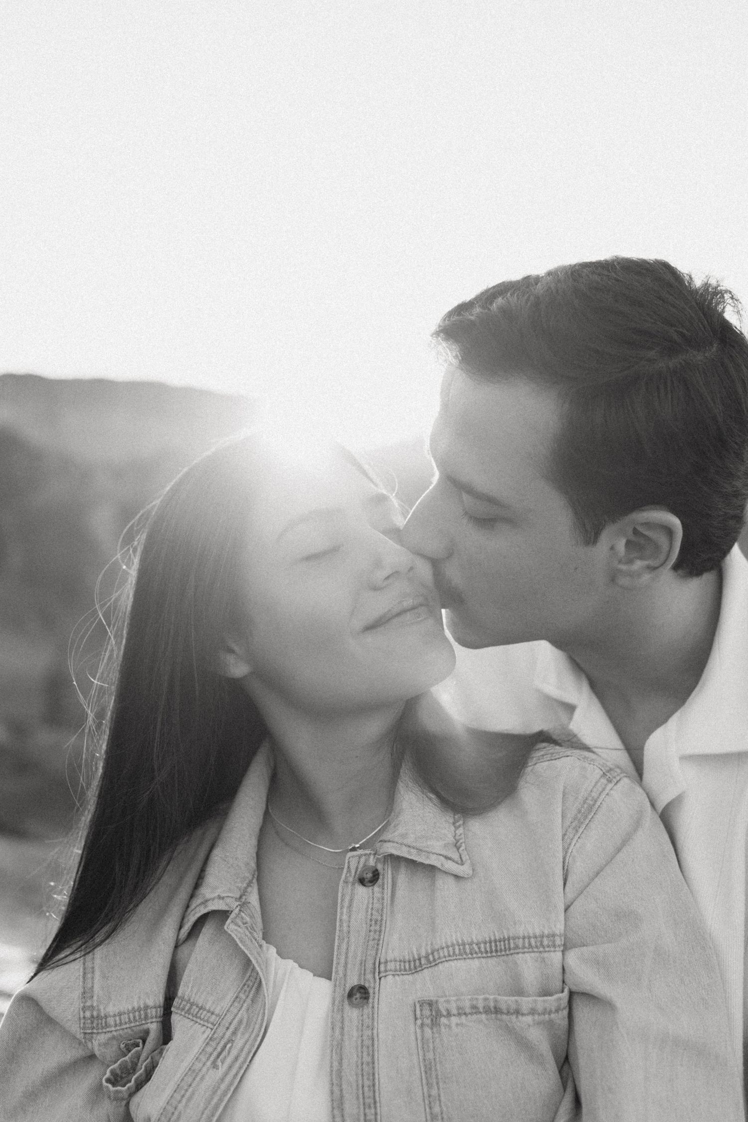Black and white photo of a man kissing the cheek of a smiling woman in a denim jacket as sunlight glows behind them.