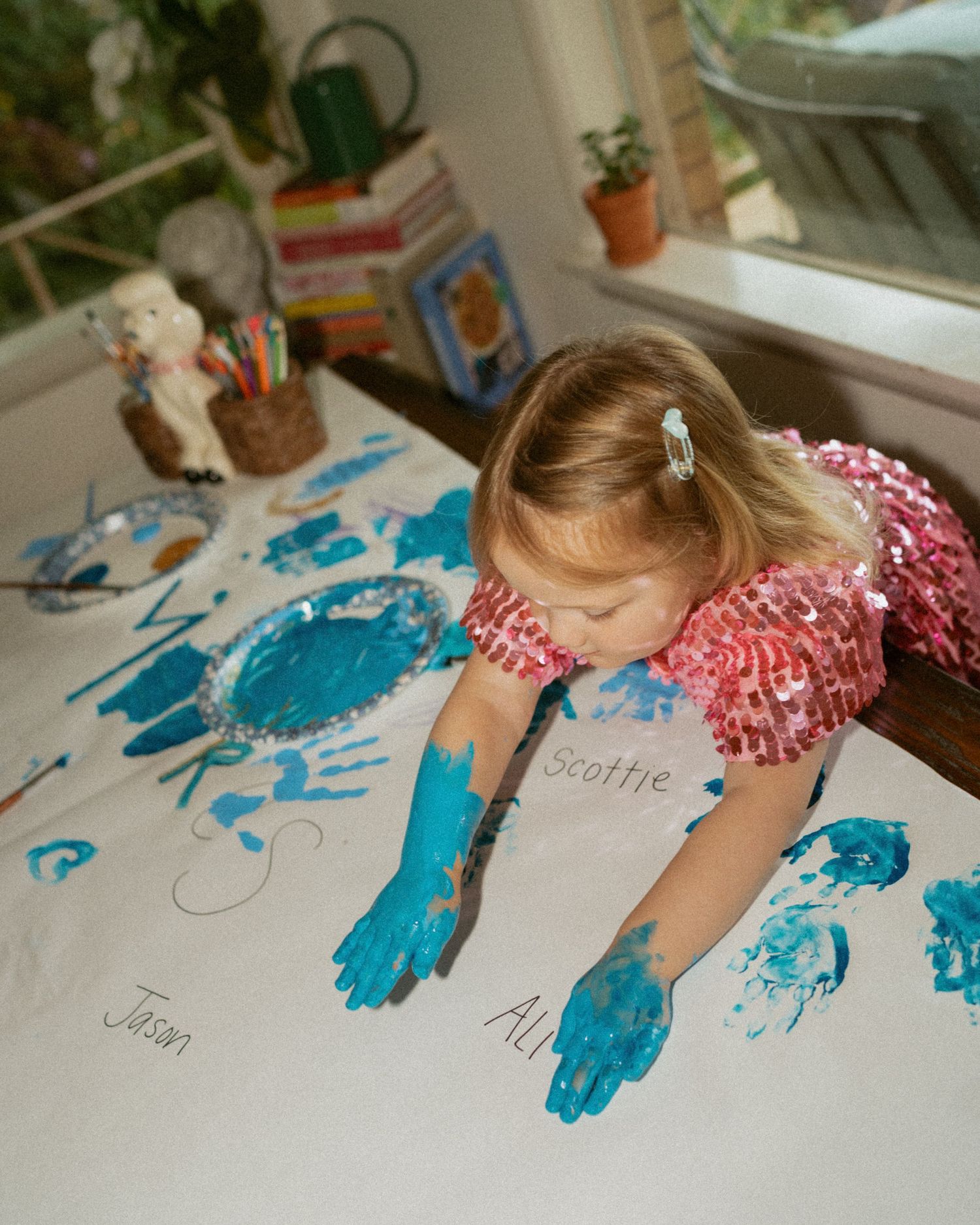 Toddler in a sparkly pink sequin dress makes blue handprints with paint on a large white paper covered in names at a table.