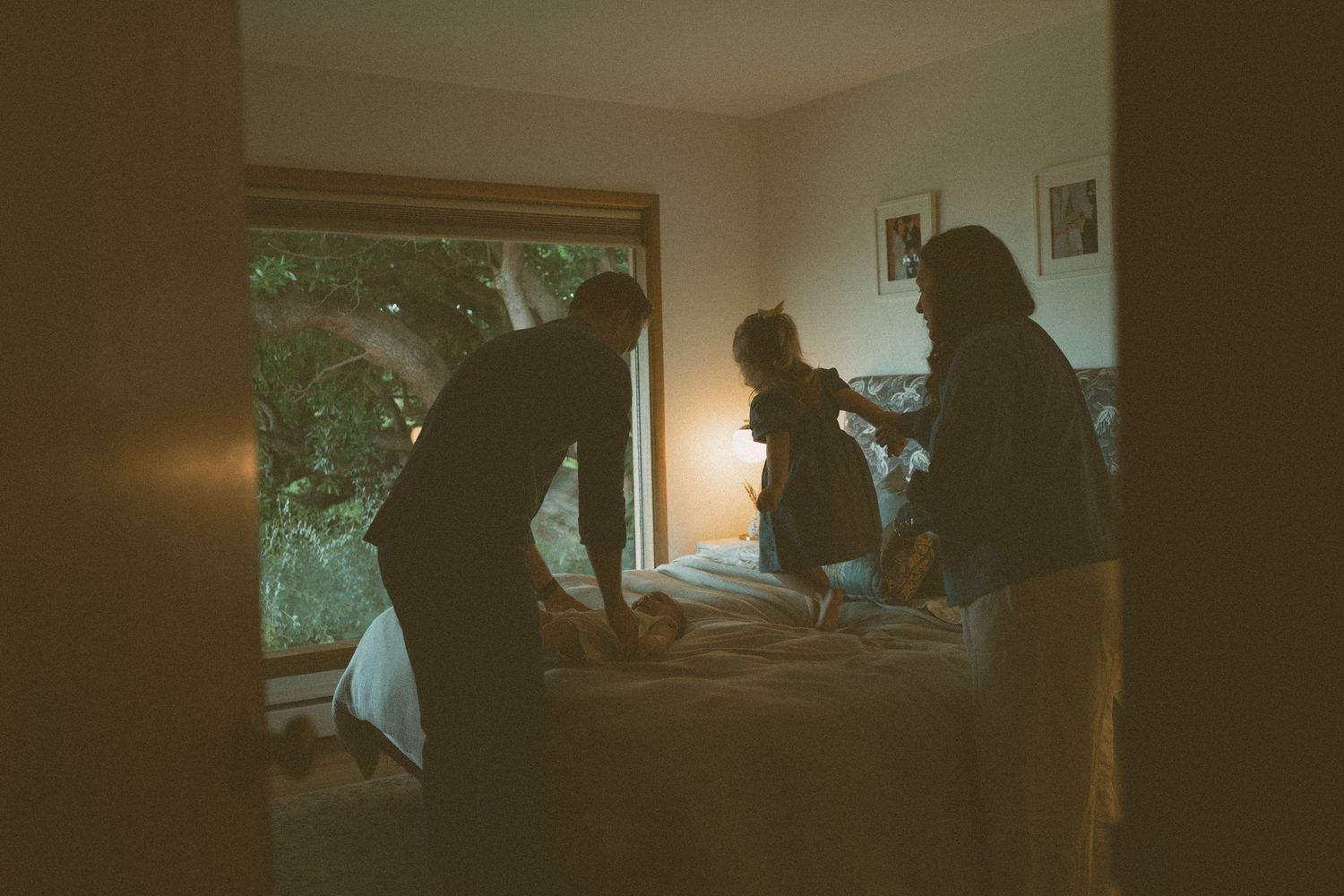 Silhouetted family of three playing on a bed in a cozy bedroom, with warm lamplight and a large window showing green trees outside.