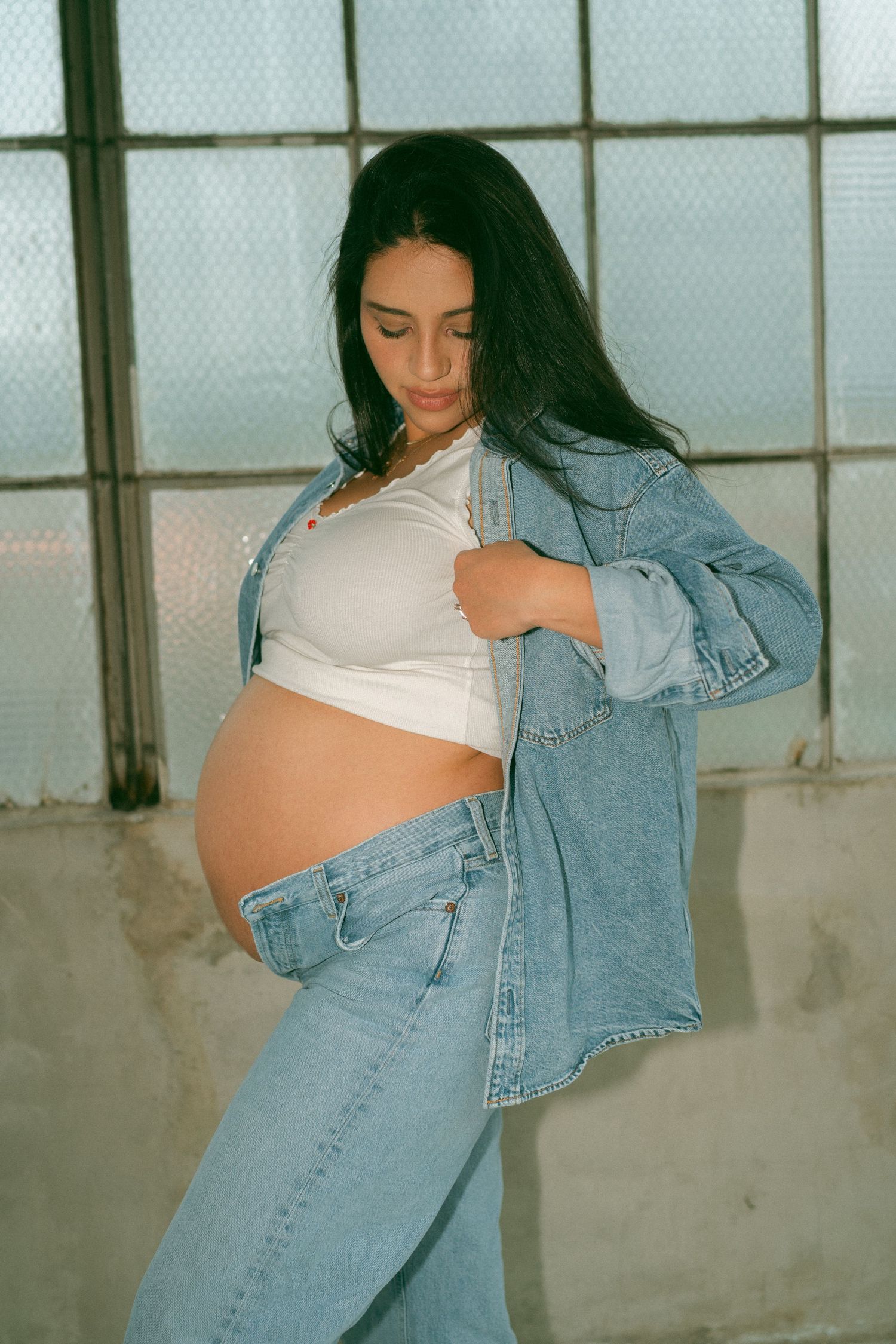 Person in denim jacket and white top posing in front of industrial windows.