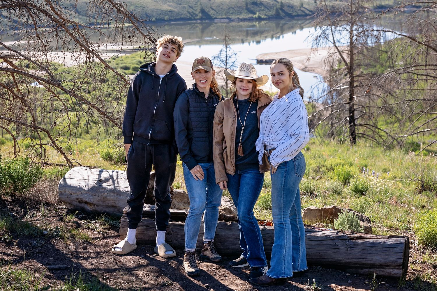 Kasia Sutton from Kasia Sutton Photography with her children  posing together outdoors near a lake and wooded hillside on a sunny day.