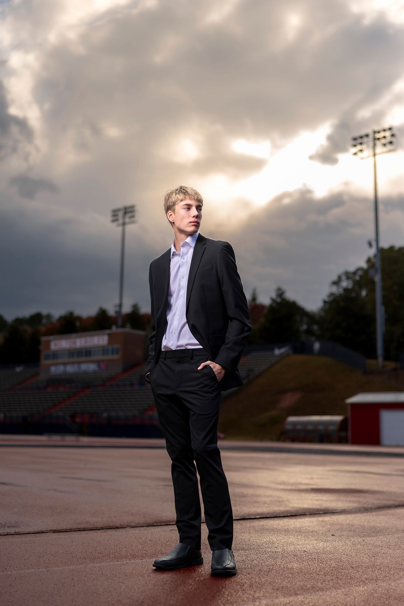 Young man in black suit stands confidently on a track field under dramatic stormy sky with stadium lights behind him.