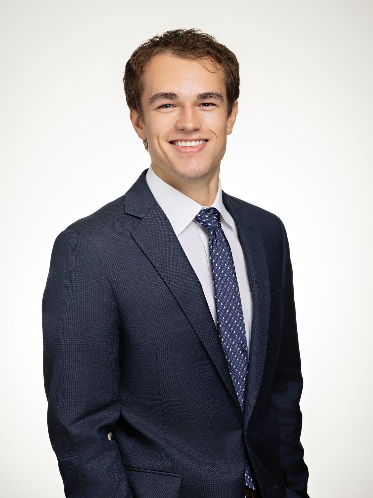 Young professional man smiling in navy suit and blue tie against white background for business headshot.