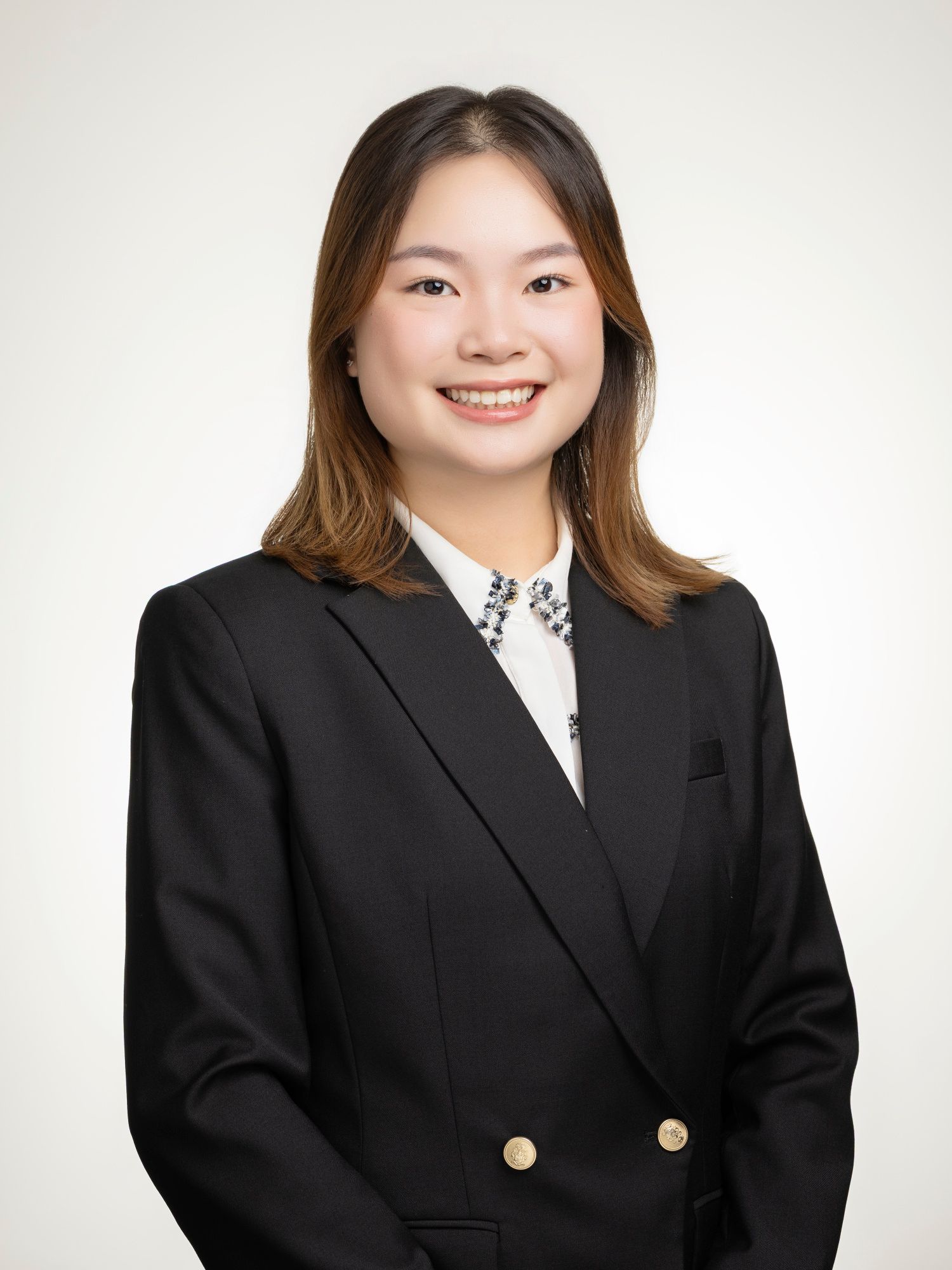 Professional headshot of a smiling young woman in a black blazer and white blouse with decorative necklace.