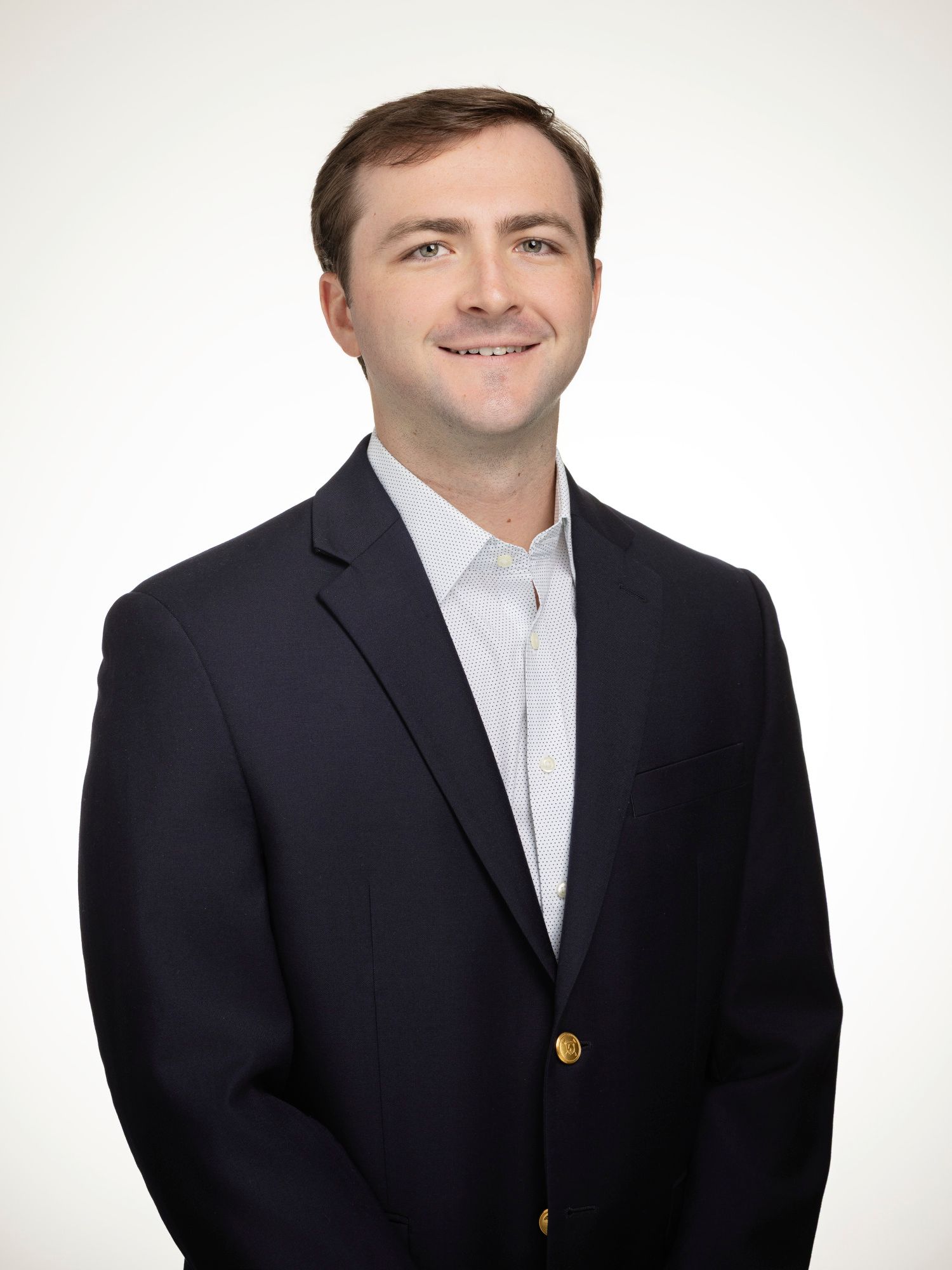 Professional headshot of a smiling young man in a navy blazer and light patterned shirt against a white background.