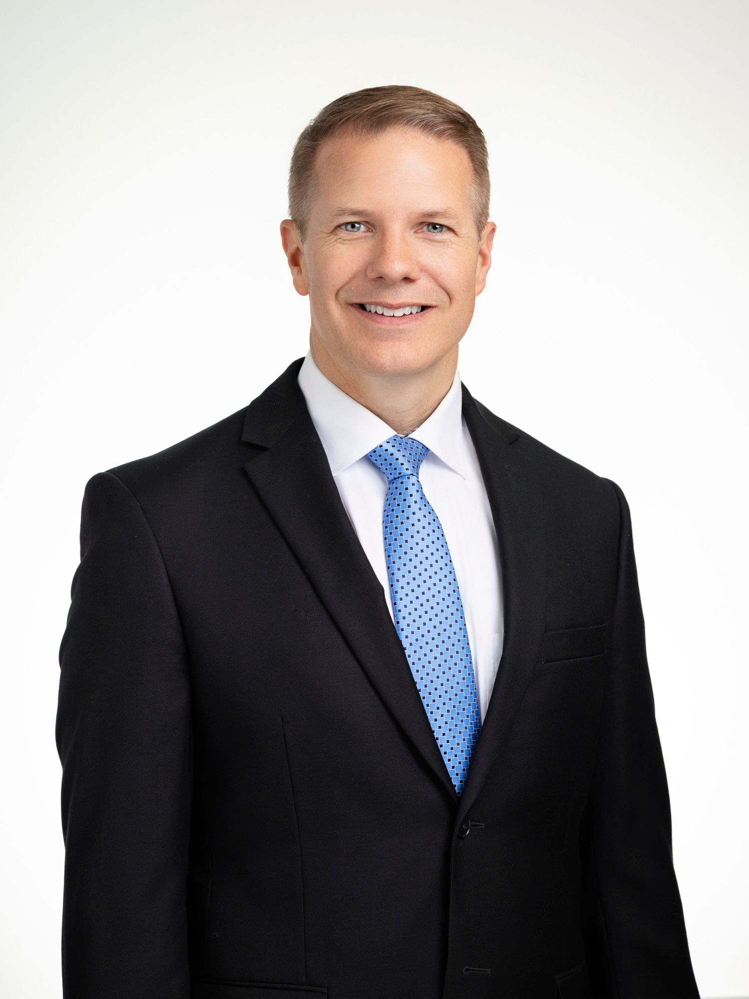 Professional headshot of a smiling man in a black suit and light blue tie against a white background.
