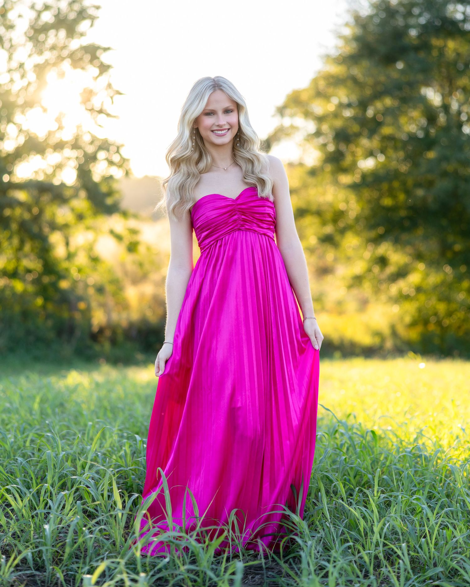 Blonde woman in a strapless hot pink maxi dress standing in a lush green field at golden hour.