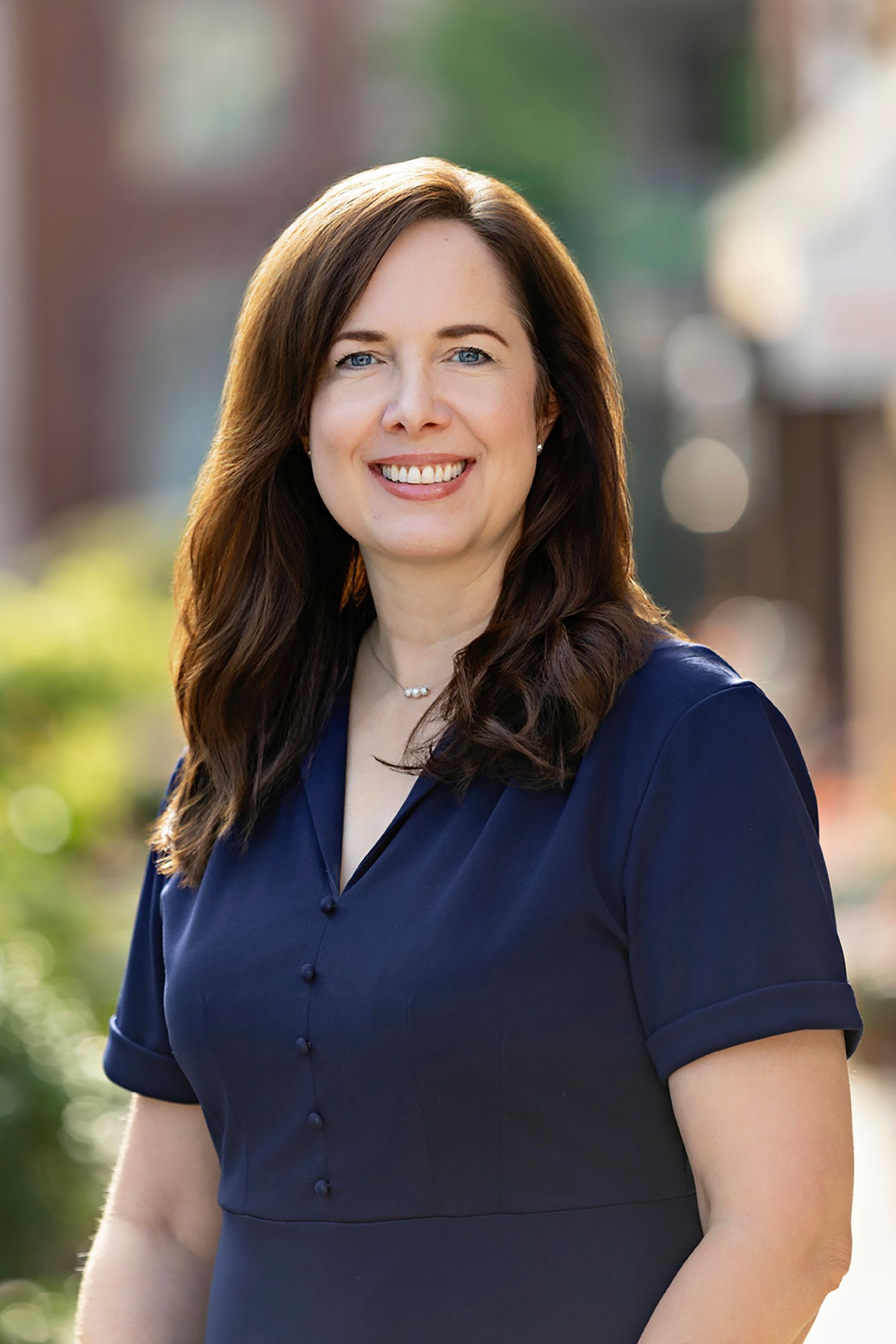 Professional woman in navy dress smiling outdoors, with long brown hair and blue eyes, in a business portrait.