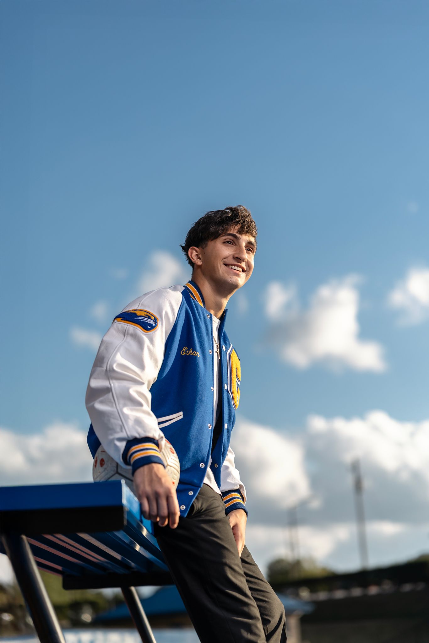 Young man in blue and white varsity jacket smiling outdoors under blue sky.