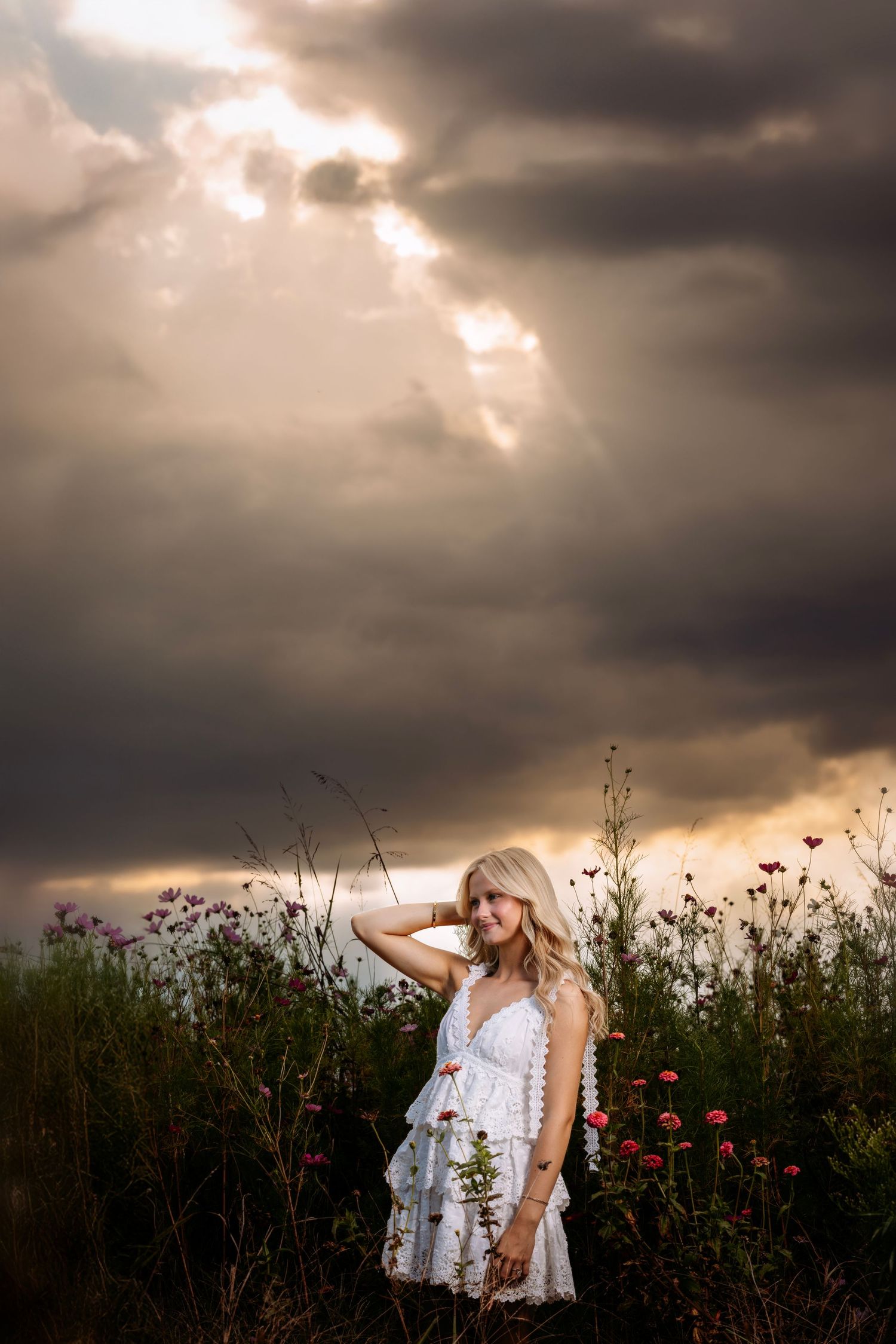 Woman in white dress standing in wildflower field under dramatic stormy sky with sunbeams breaking through clouds.