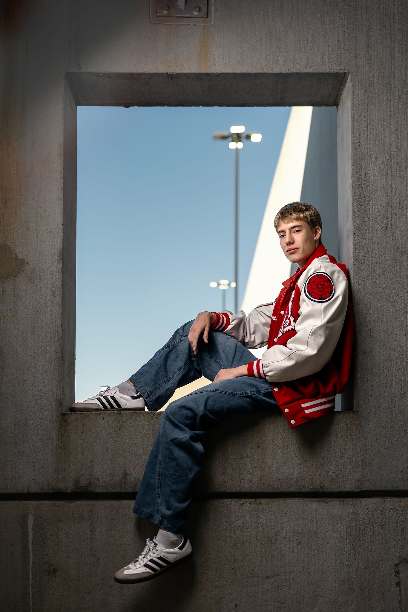Young man in red varsity jacket and jeans sitting in a concrete window frame against a blue sky.