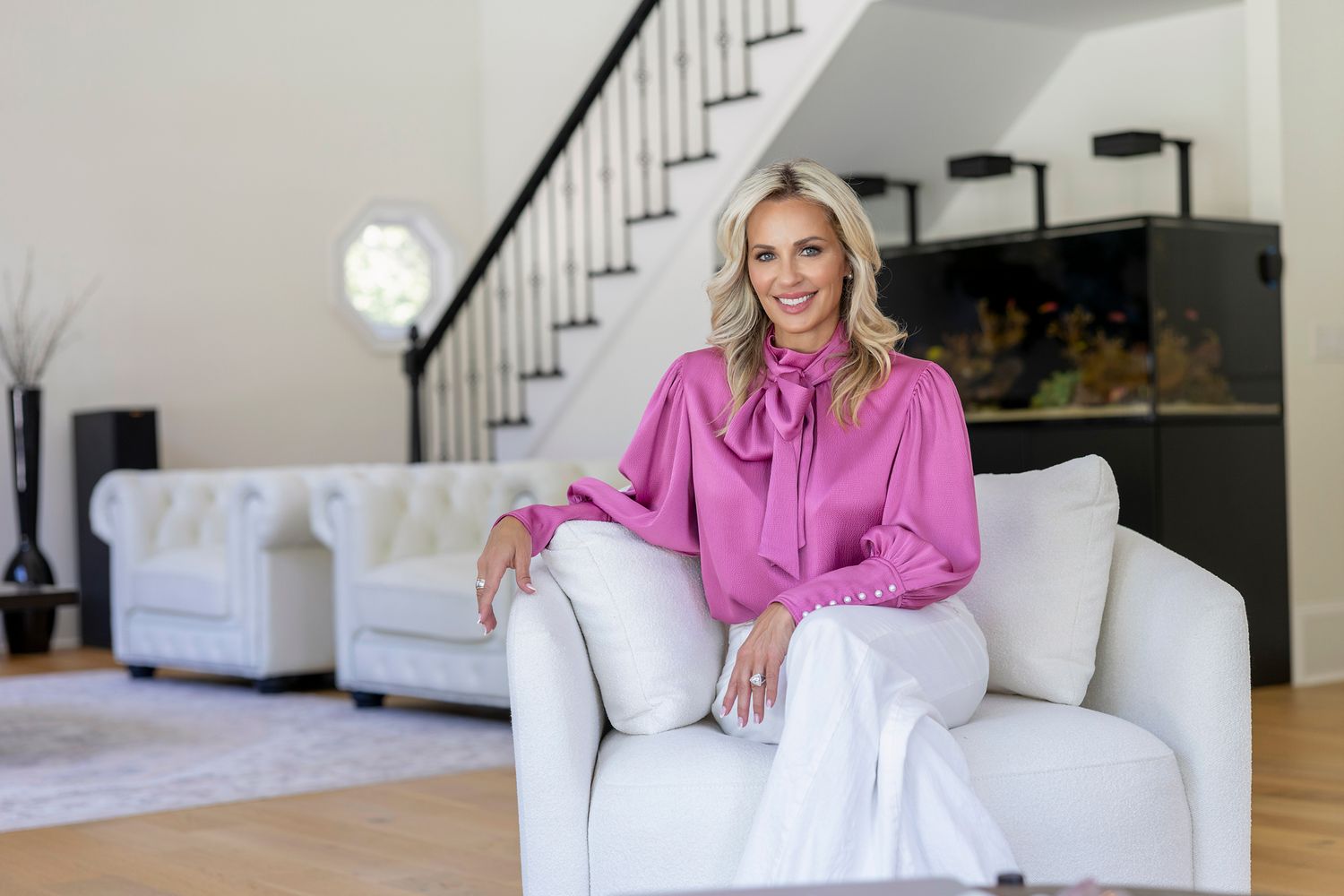 Smiling blonde woman in pink blouse and white pants sitting on white sofa in modern living room.