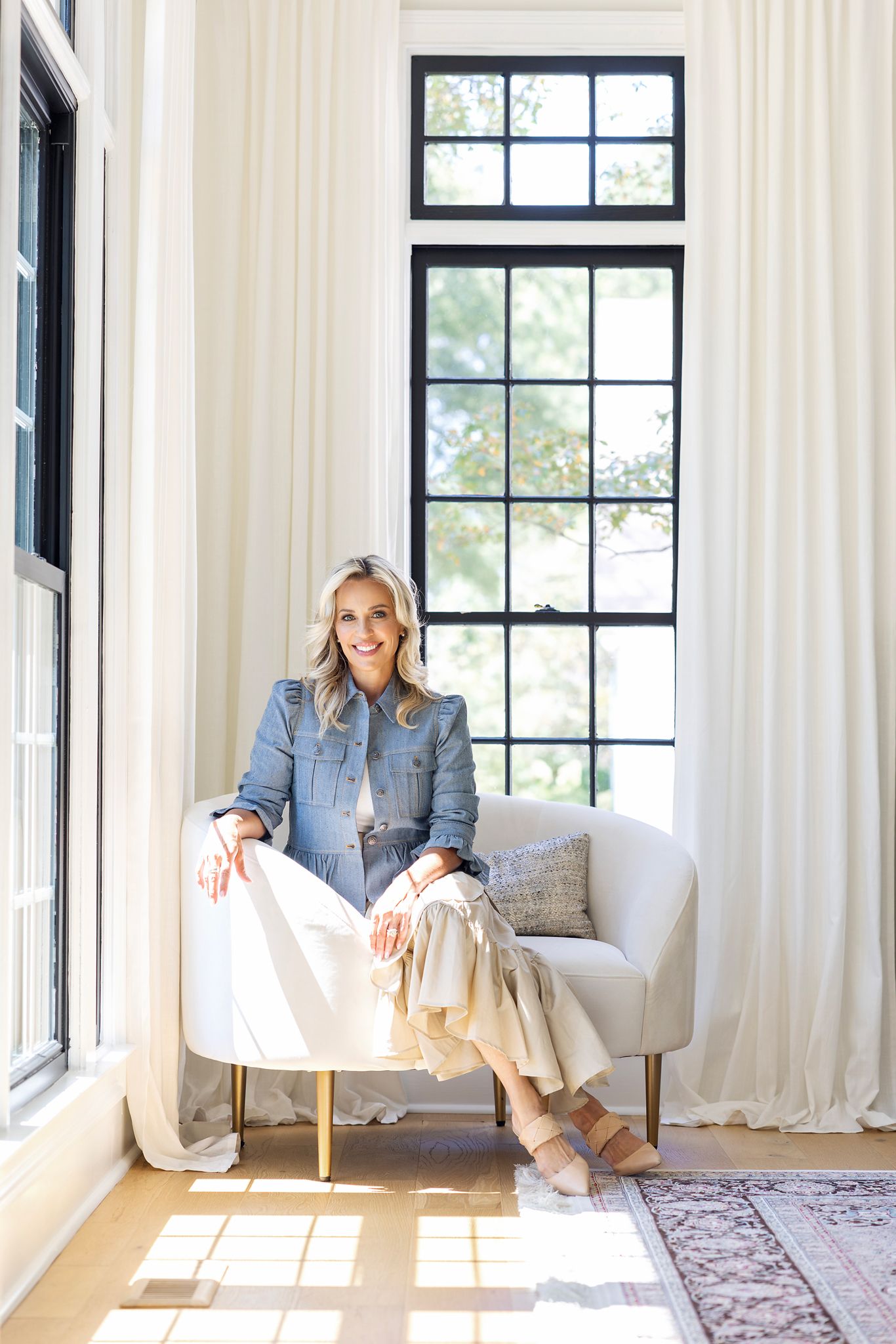 Smiling blonde woman in denim jacket seated on white chair by large black-framed windows with white curtains.