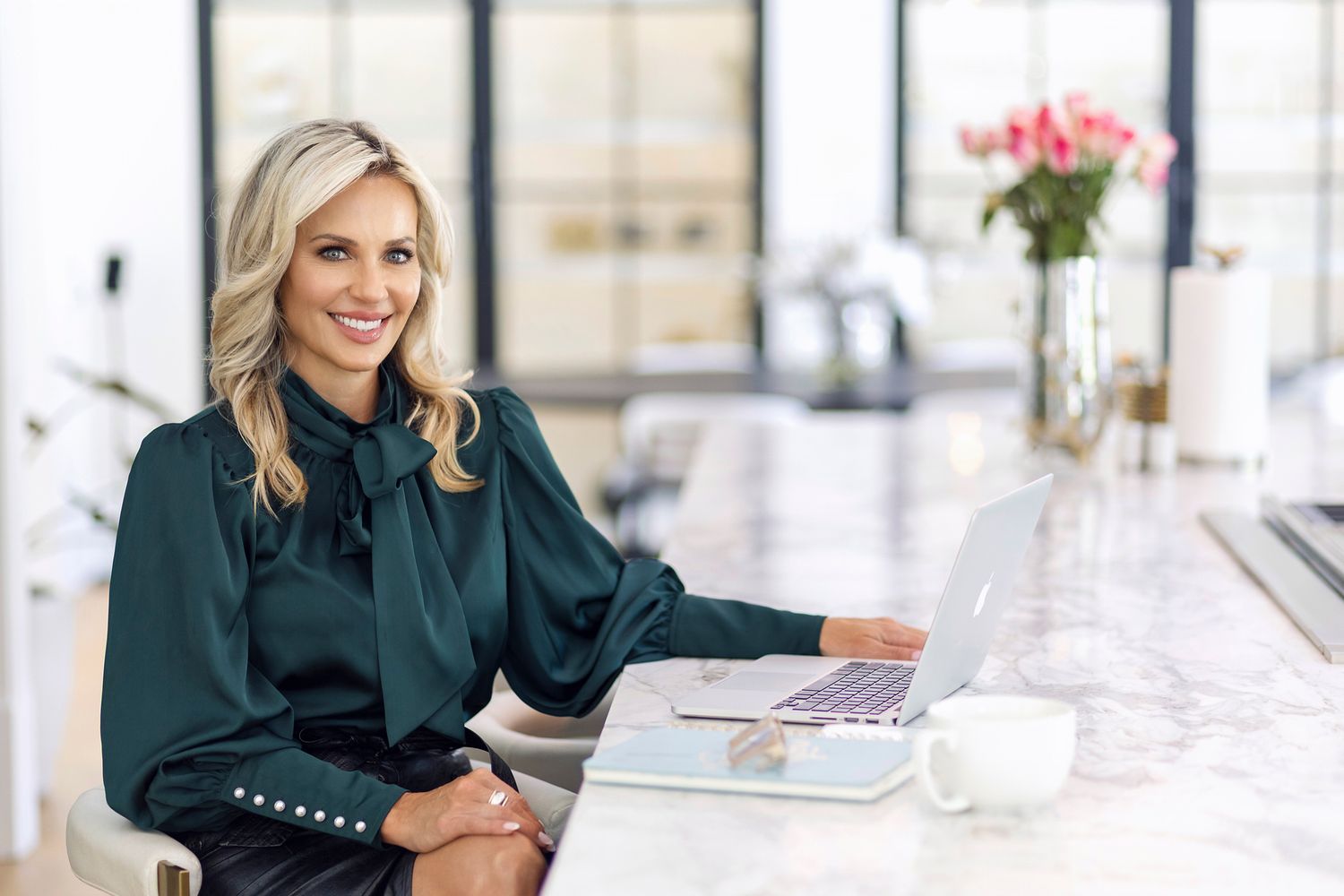 Smiling blonde businesswoman in green blouse working on laptop at marble desk in bright modern office.