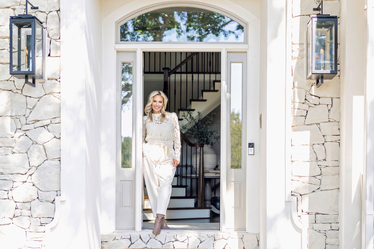 Elegant woman in white outfit standing in doorway of luxury stone home with black lanterns and grand staircase.