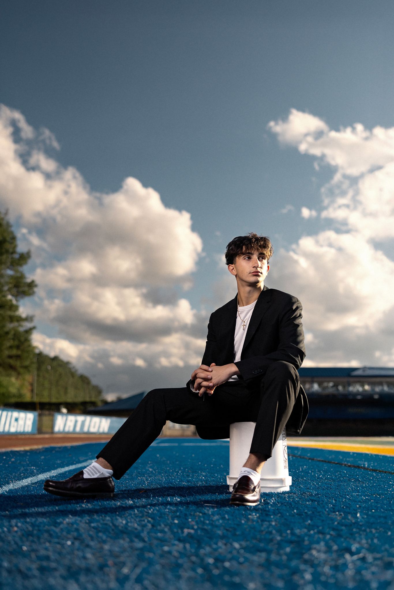 Young man in black suit sitting on track at outdoor stadium under dramatic cloudy blue sky.
