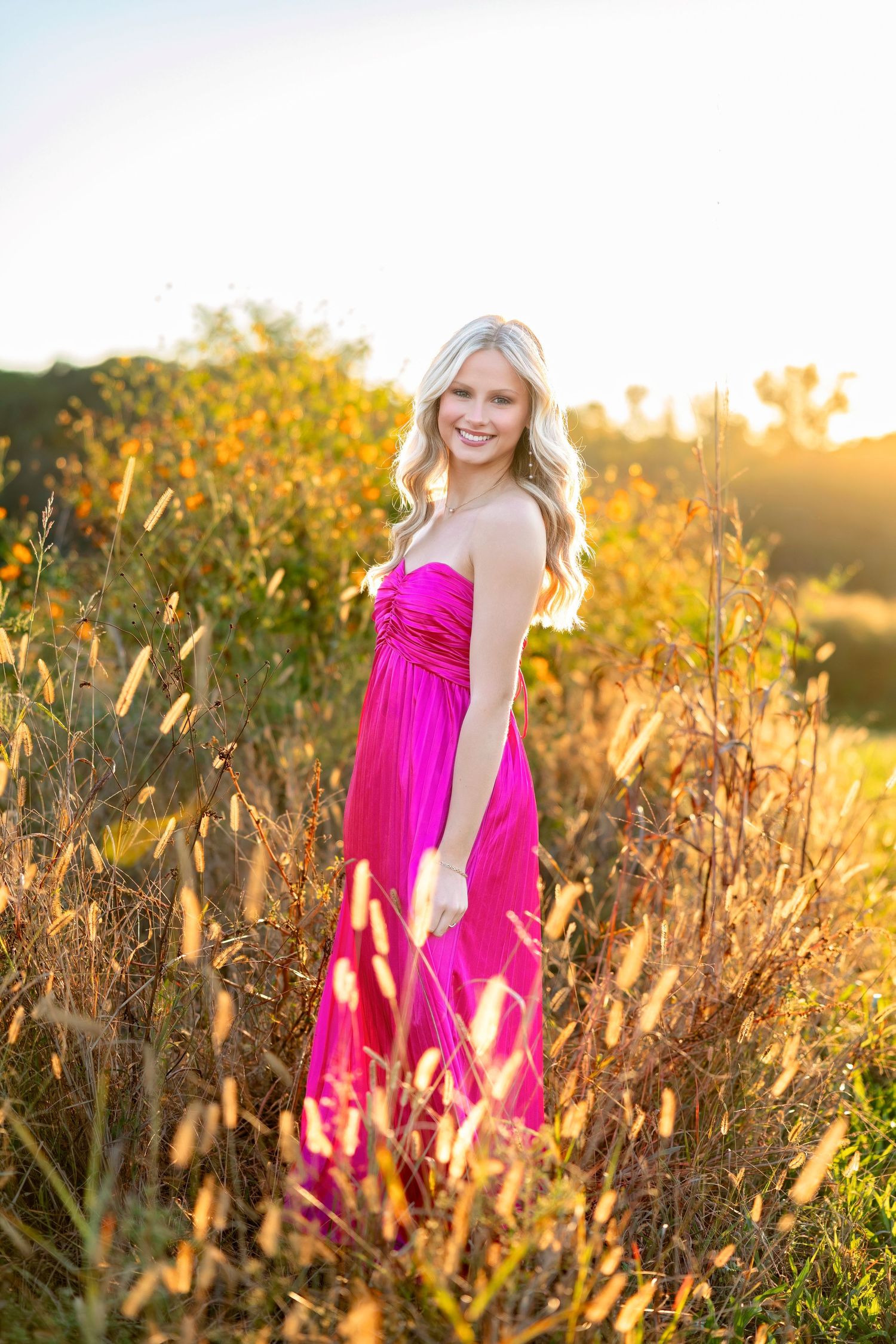 Smiling blonde woman in pink strapless maxi dress standing in golden sunlit field with wildflowers.