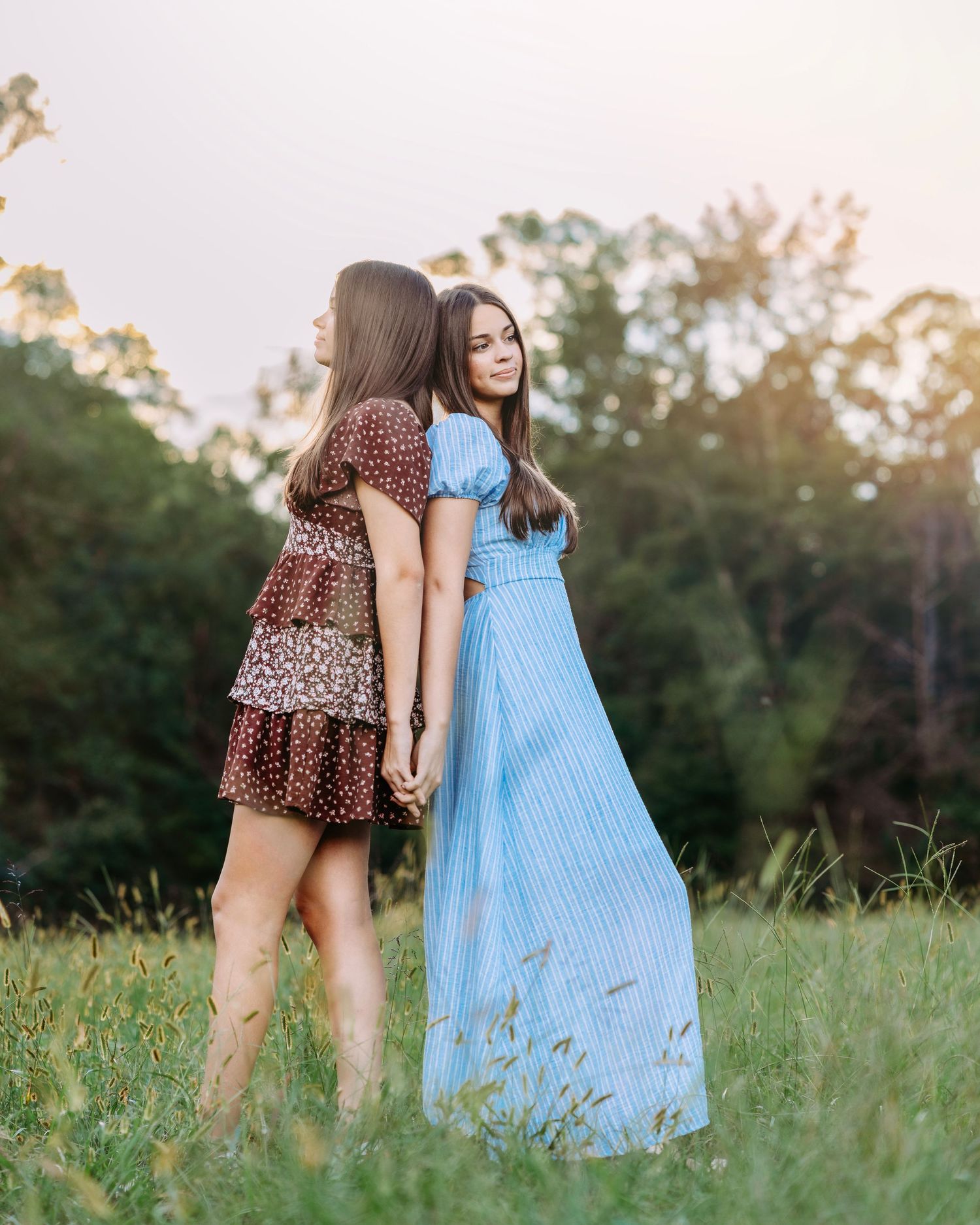 Two girls in summer dresses hold hands in a sunlit meadow, one in blue maxi dress, other in brown polka dot mini.
