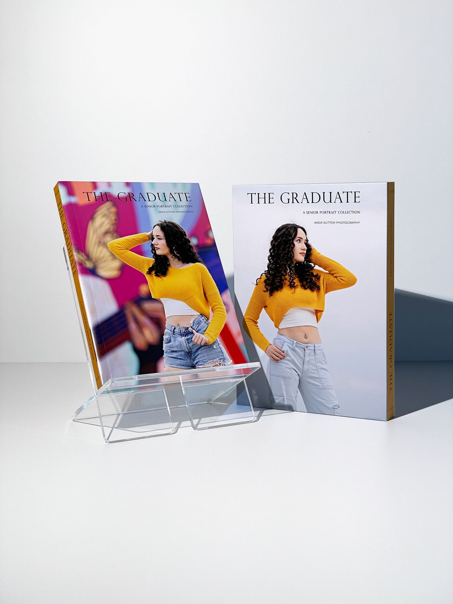 Two copies of The Graduate magazine featuring a woman in a yellow crop top and jeans, displayed on a clear acrylic stand.