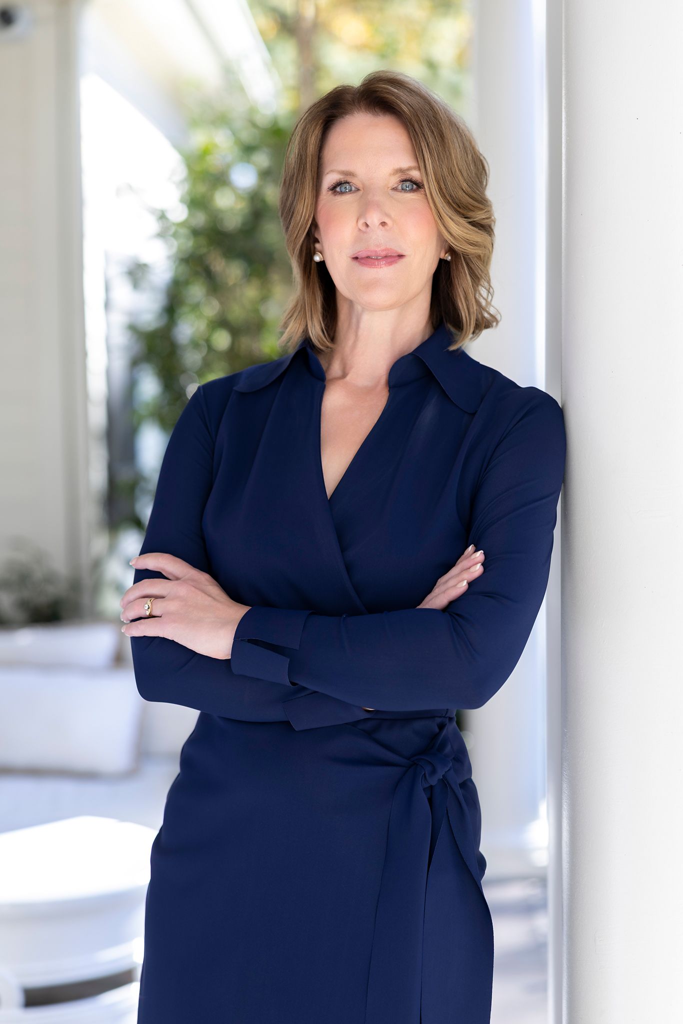 Professional woman in navy wrap dress with arms crossed, leaning against white column in bright modern setting.