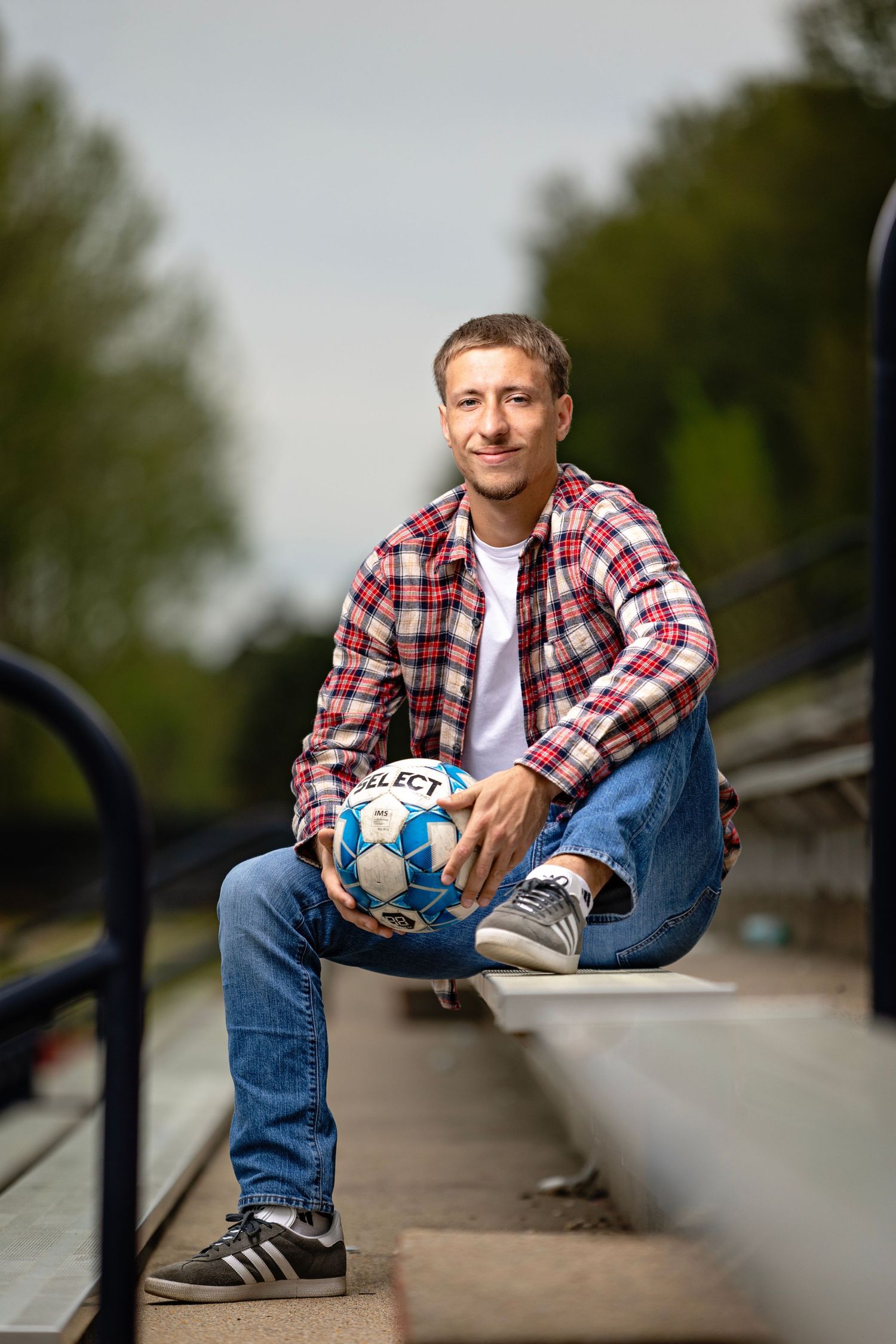 Senior boy in plaid shirt and jeans sitting on bleachers holding a Select soccer ball outdoors.