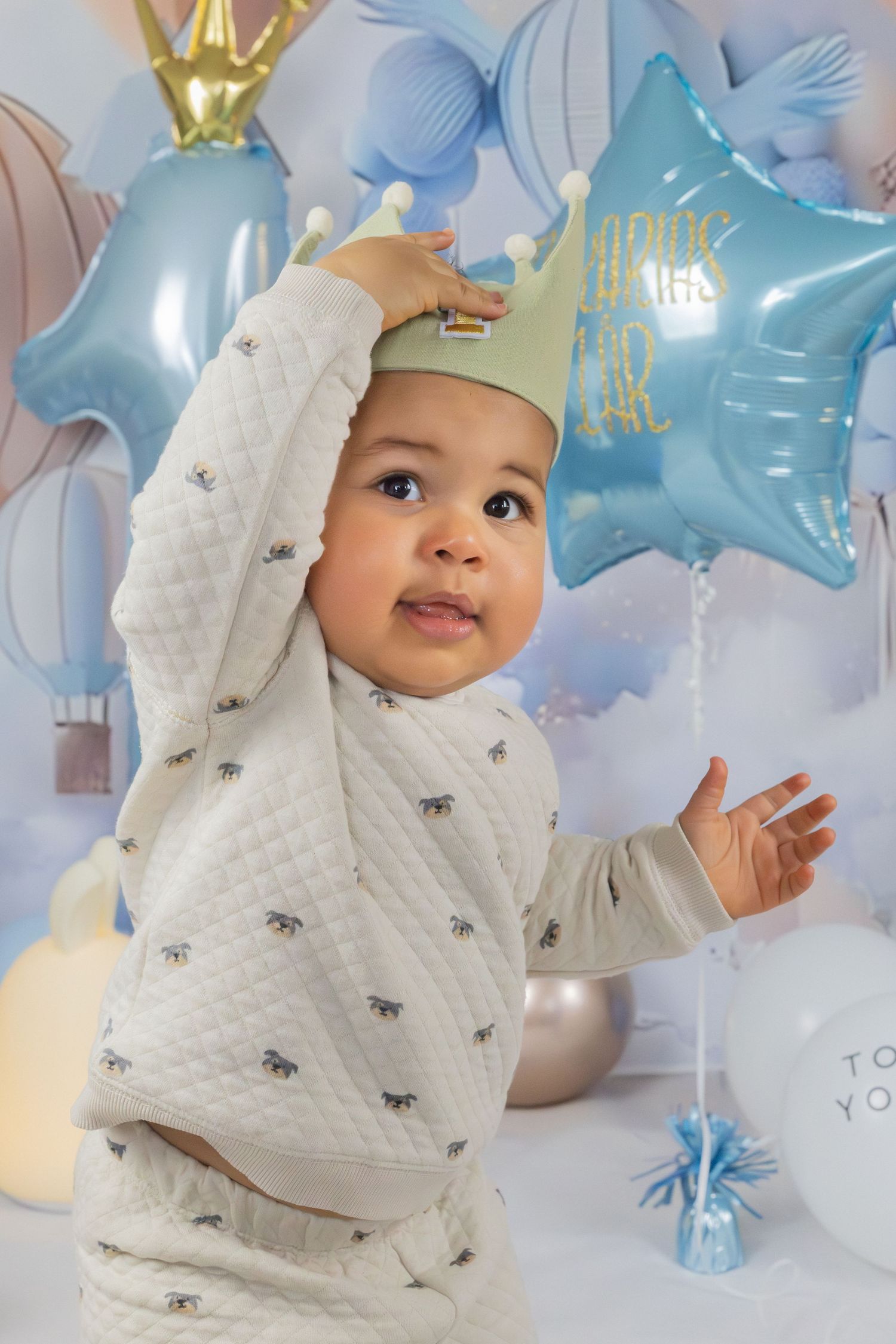 Barn i ljus outfit sträcker sig mot blå ballonger inomhus, porträttfoto med ballongbakgrund. Födelsedagsfotografering.
