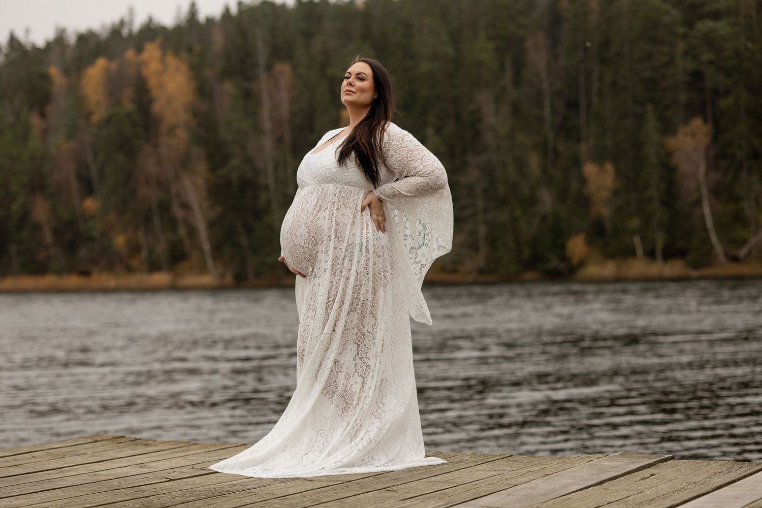 Gravid kvinna i vit spetsklänning poserar vid en sjö med höstskog i bakgrunden – gravidfotografering utomhus i naturen.