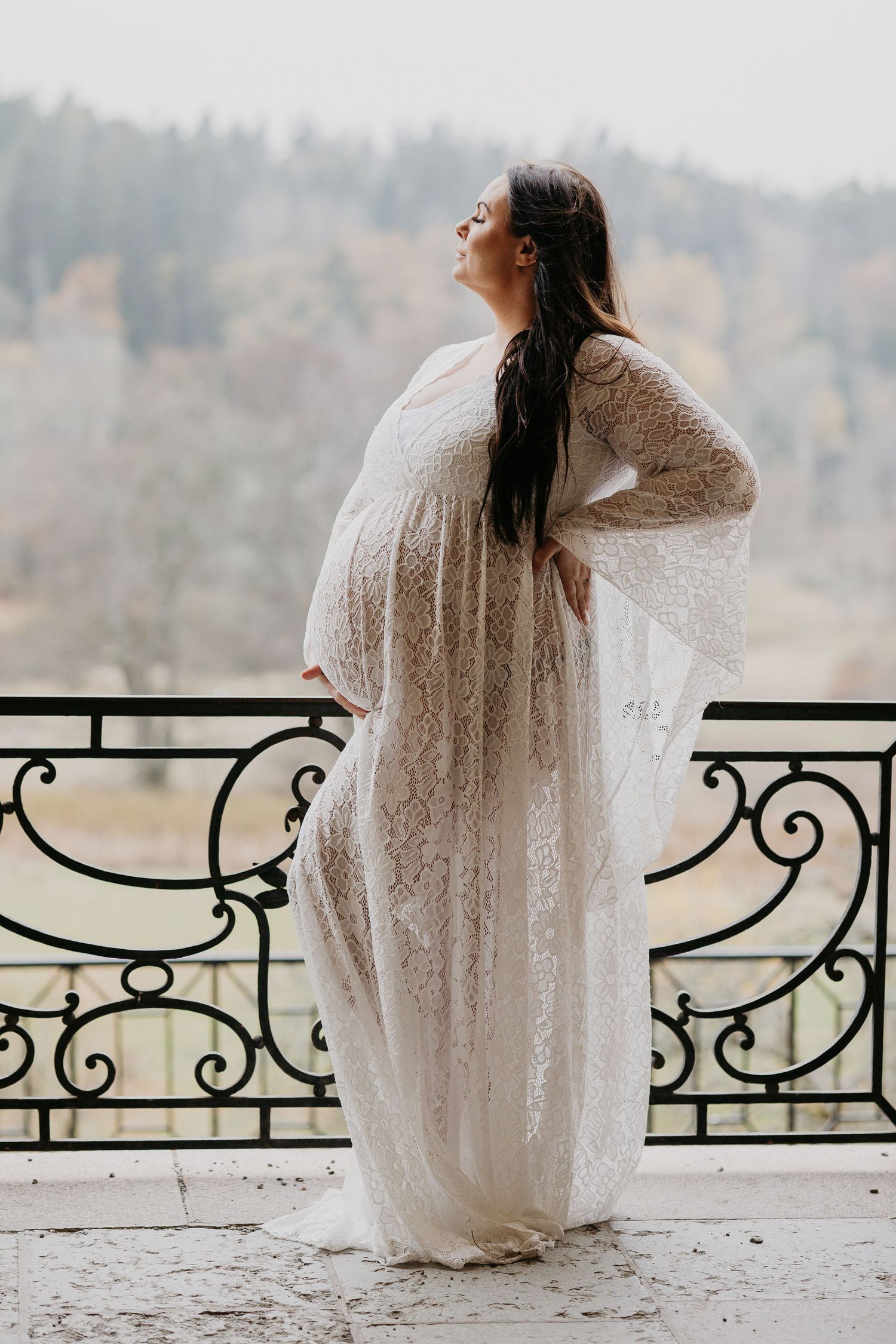 Gravid kvinna i vit spetsklänning poserar mjukt på balkong med dekorativa räcken under gravidfotografering.