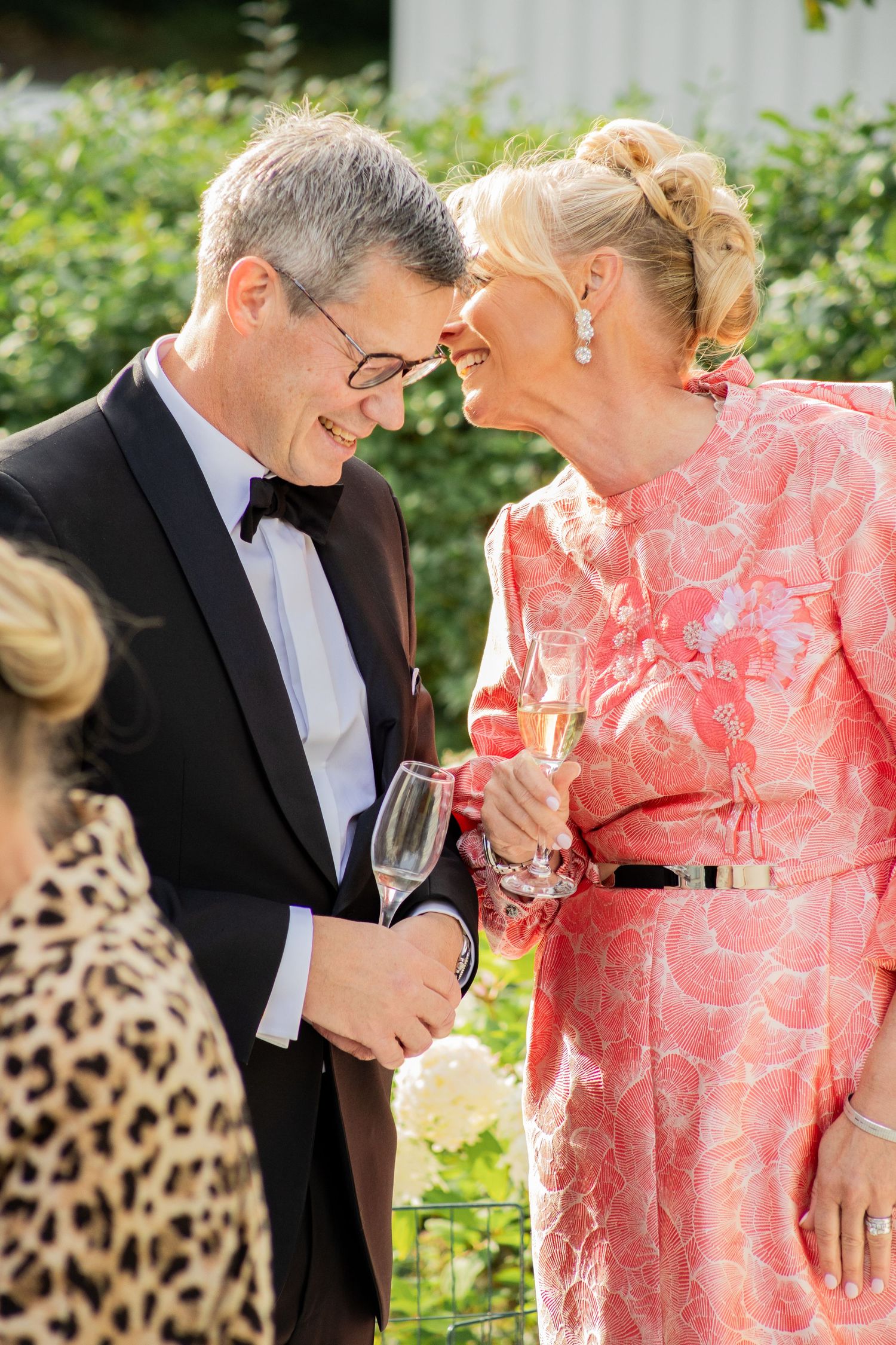 Äldre par i festkläder håller champagneglas och samtalar utomhus, bröllopsmingel i trädgård.