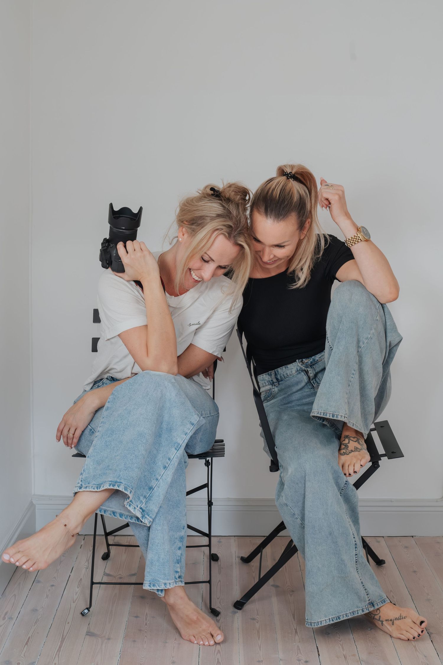 Två fotografer i ljusa toppar och blå jeans sitter på stol med kameror, porträtt av fotografduo.