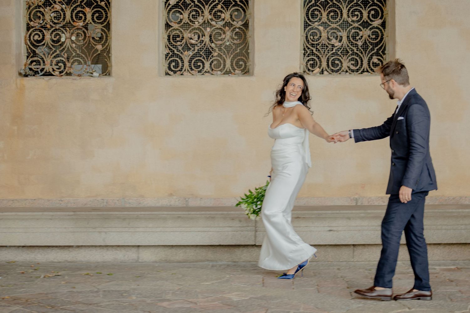 Bride in white strapless gown holds groom's hand in navy suit, dancing outdoors near ornate iron window.
