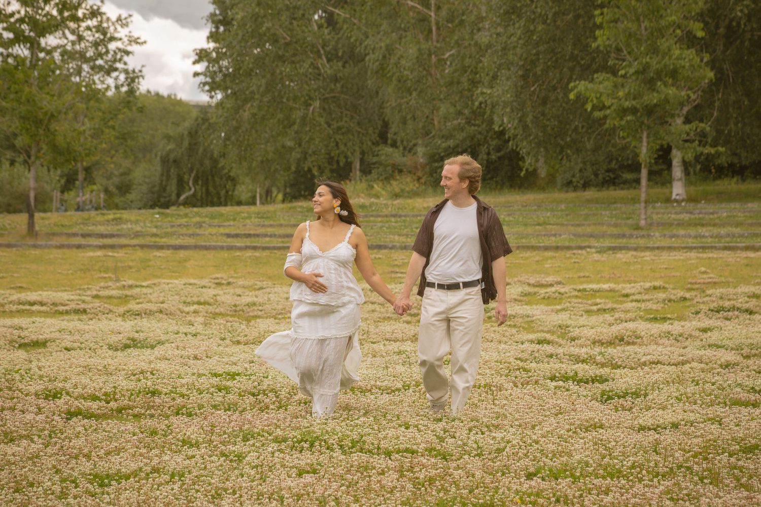 Couple holding hands walking through a blooming white flower field with lush green trees in the background.