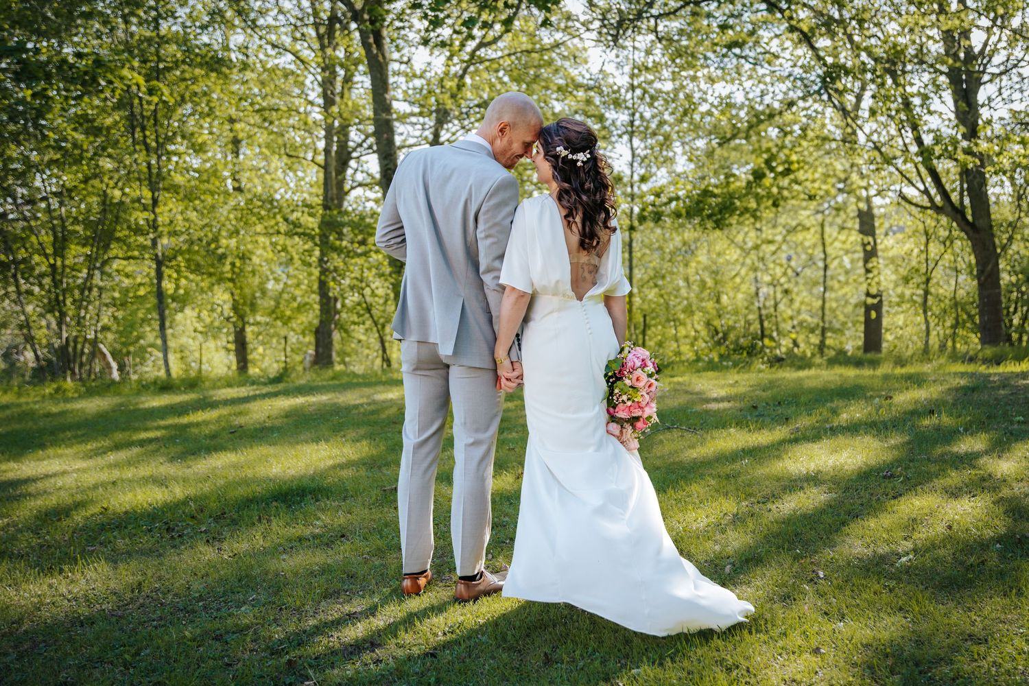 Brudpar håller om varandra i grön sommaräng under utomhusfotografering med träd i bakgrunden.