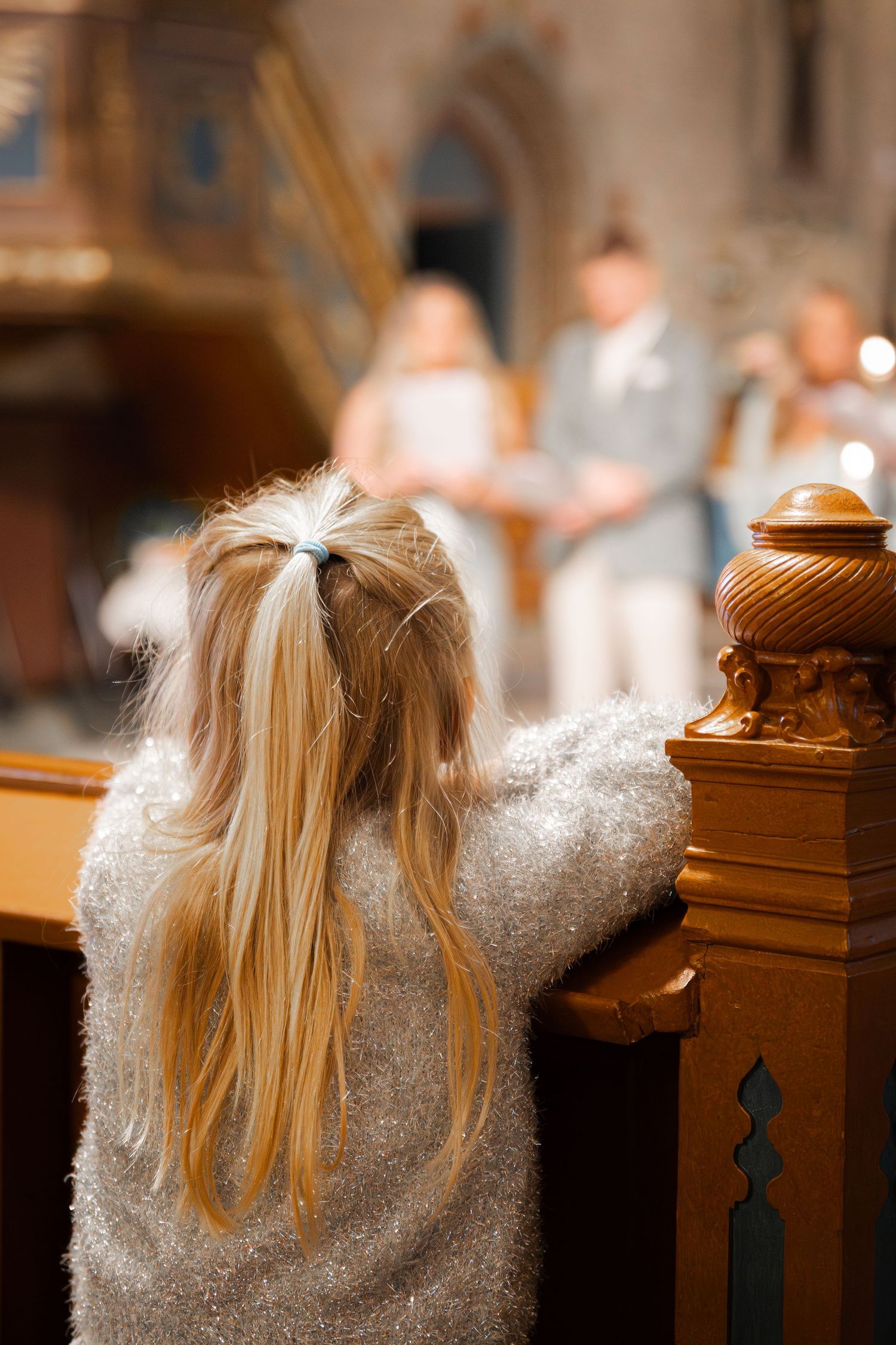 Litet barn med blond hästsvans lutar sig mot kyrkbänk och tittar mot dopet i bakgrunden, stämningsbild från dop i kyrka.