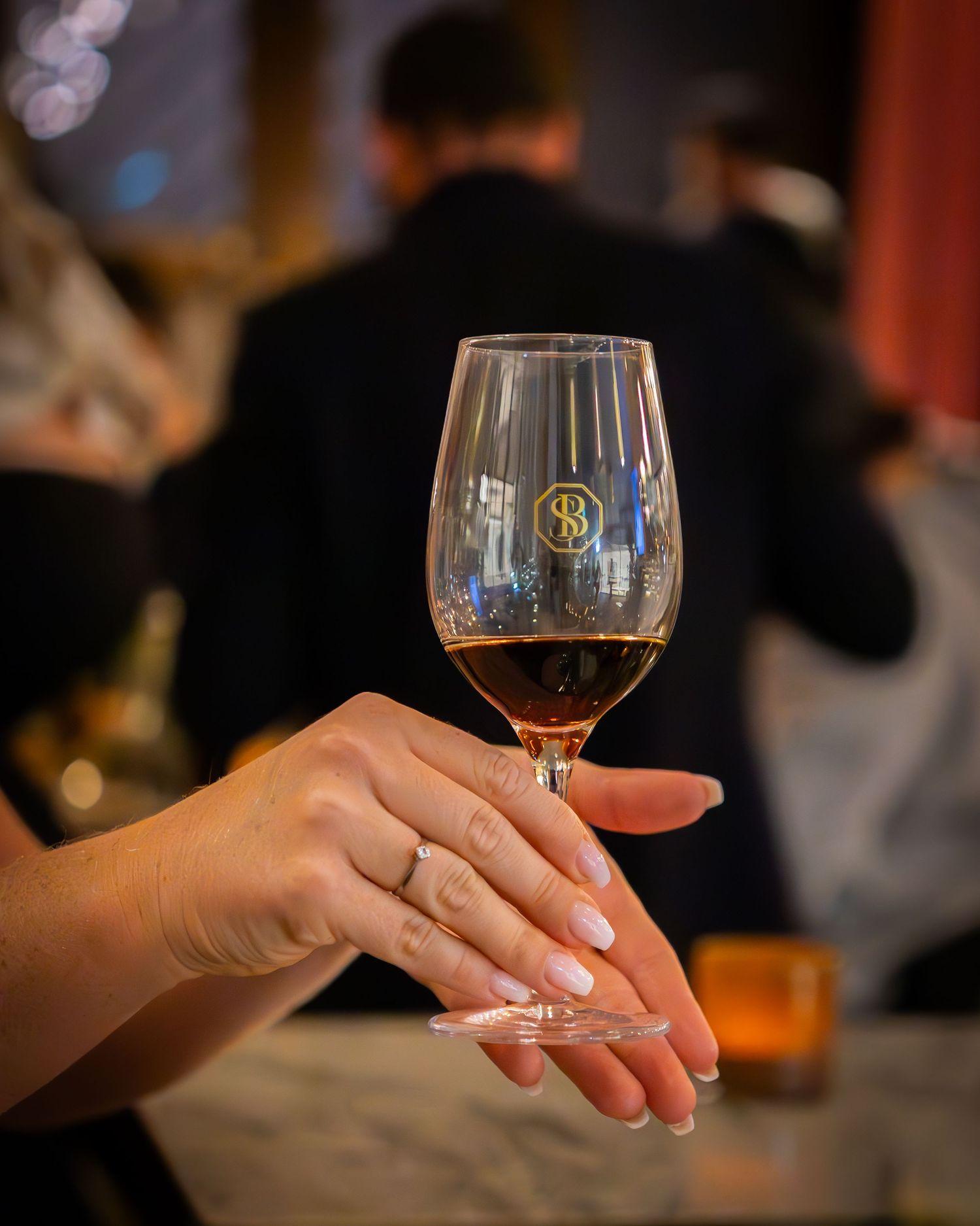 Hand med vinglas i fokus under vinprovning, med vacker ring och suddig bakgrund av minglande gäster.