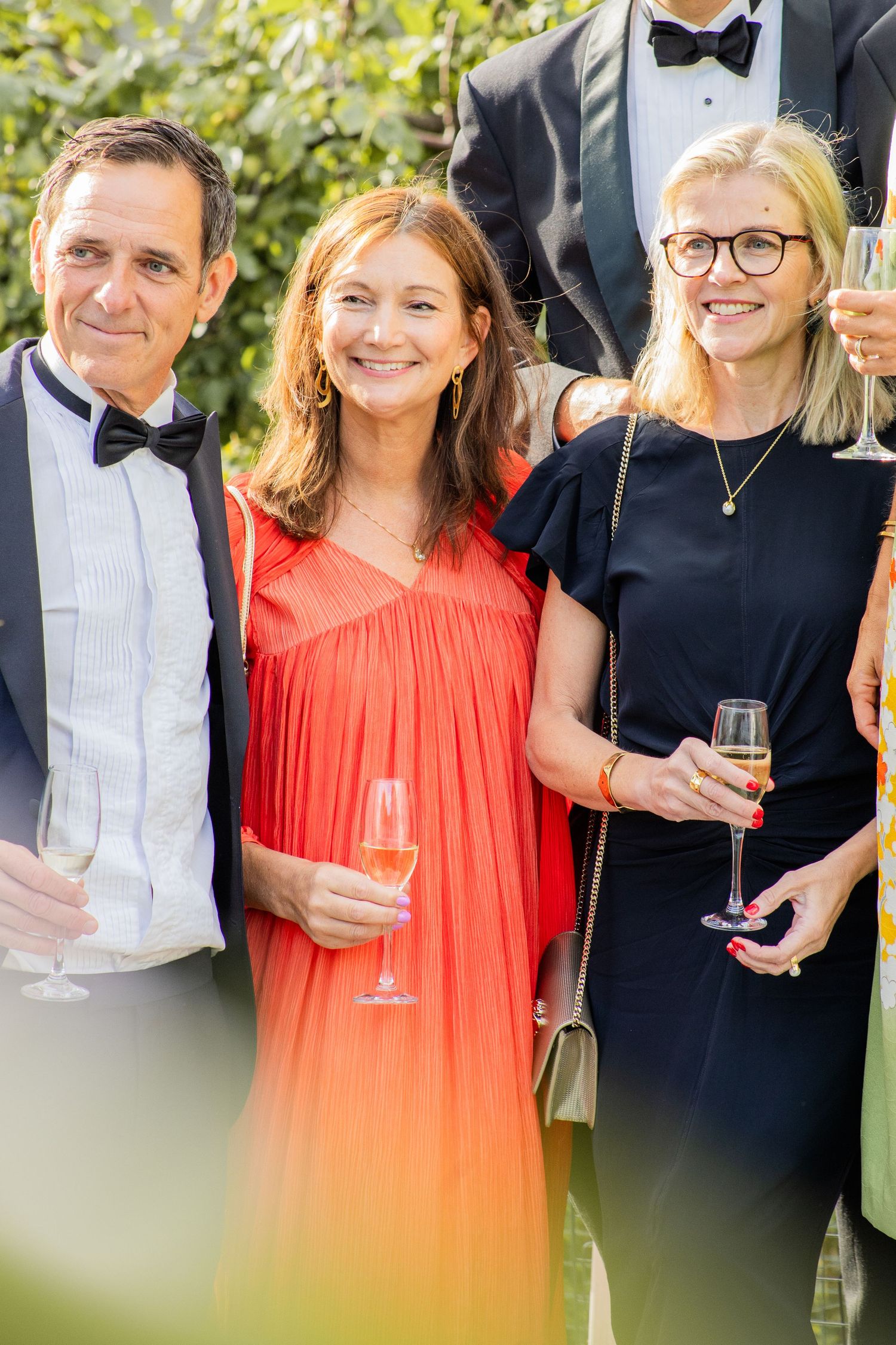 Tre gäster i festkläder håller champagneglas utomhus, mingel på bröllop eller sommarevent.